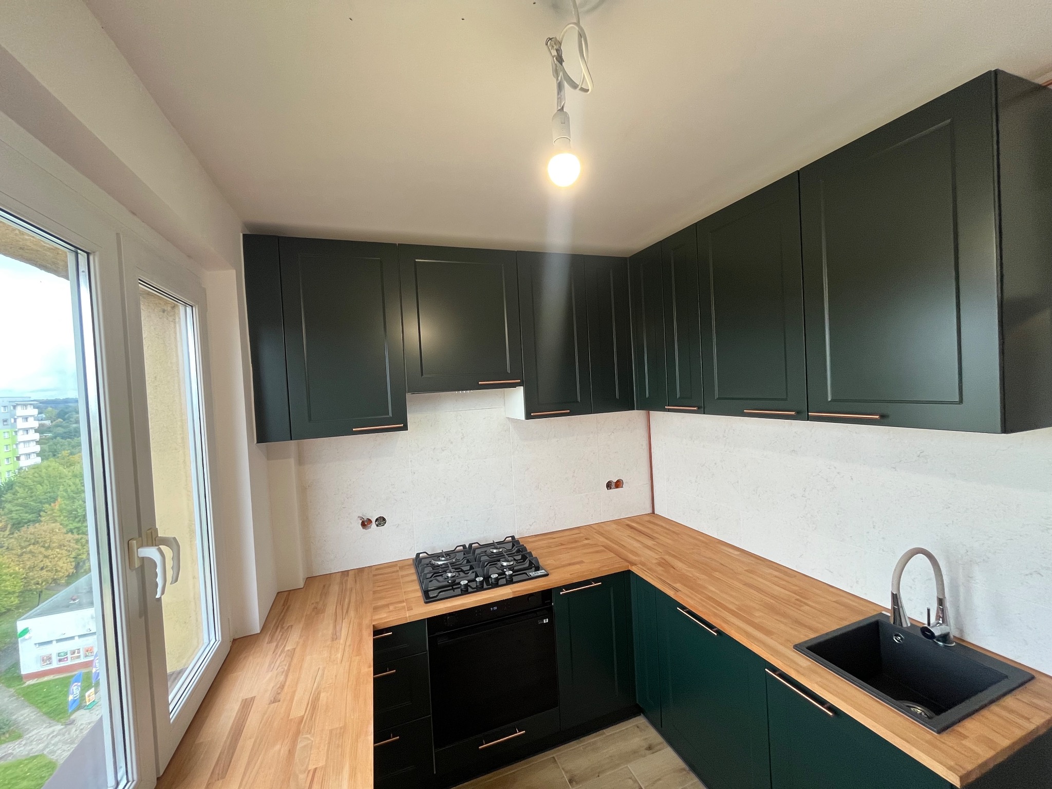 Nowoczesna kuchnia w odcieniach zieleni i drewna z widokiem z okna. Uwagę przykuwają ciemnozielone szafki z miedzianymi uchwytami i drewniany blat.