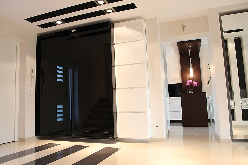 Nowoczesna szafa przesuwna z czarnymi, błyszczącymi frontami i białymi panelami bocznymi w przestronnym holu z czarno-białą podłogą i dekoracyjnym oświetleniem.