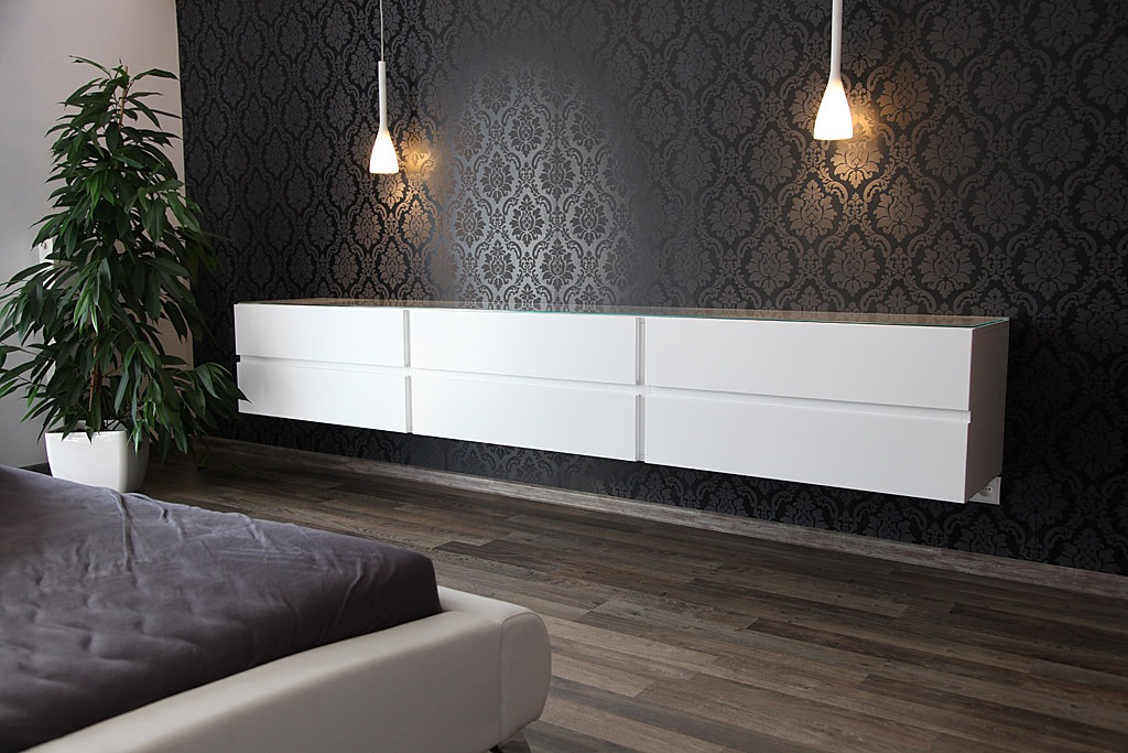 Biała, wisząca komoda z sześcioma szufladami, z blatem ze szkła, na tle ściany w ciemne, ozdobne wzory. Nad komodą wiszą dwie lampy. W tle widoczny kwiat doniczkowy.