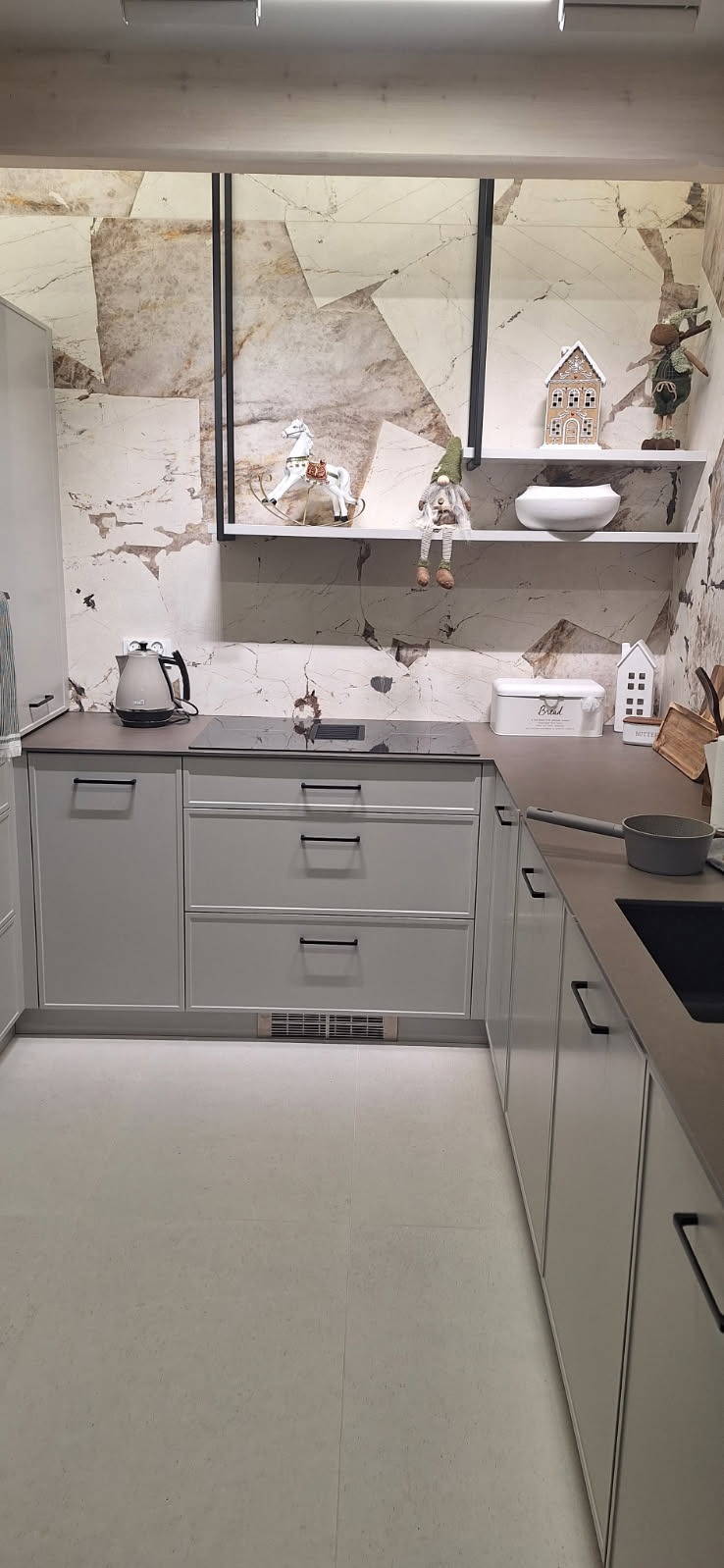 Jasna kuchnia z szarymi szafkami, czarnymi uchwytami i blatem. Marmurowa ściana z półkami dekorowanymi figurkami i oświetleniem LED.