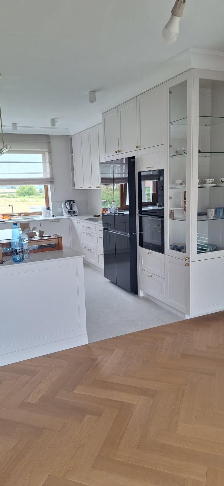 Jasna kuchnia na wymiar z czarną lodówką side-by-side i szklaną witryną. Parkiet w jodełkę na podłodze. Widok na zewnątrz przez okno.