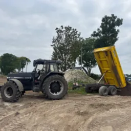 Czarny traktor Case 5150 z żółtą przyczepą wywrotką, wysypujący ziemię na placu budowy. W tle drzewa i domy.