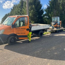 GRAC-KOP Gracjan Budzelewski - Pomarańczowa ciężarówka z przyczepą i małą koparką, pracownik w odblaskowym ubraniu na tle drzew. Napis na ciężarówce: Prace Ziemne, usługi koparko-ładowarką.