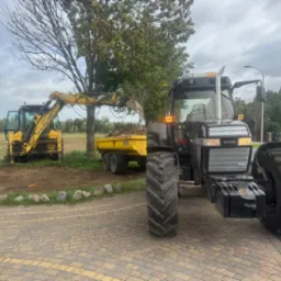 Żółta koparka ładuje ziemię na przyczepę ciągniętą przez traktor na tle drzew i pola. Pracownik z grabiami wyrównuje teren. Widok z kostki brukowej.