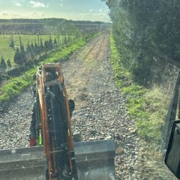 GRAC-KOP Gracjan Budzelewski - Widok z kabiny koparki na drogę z tłucznia, otoczoną zielenią i drzewami. W tle widoczne pole i niebo. Perspektywa z wnętrza maszyny.