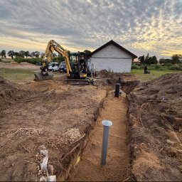 GRAC-KOP Gracjan Budzelewski - Żółta mini-koparka kopie głęboki rów pod instalacje, z widocznymi rurami, na tle budowanego domu z białej cegły i zachmurzonego nieba o zachodzie słońca.