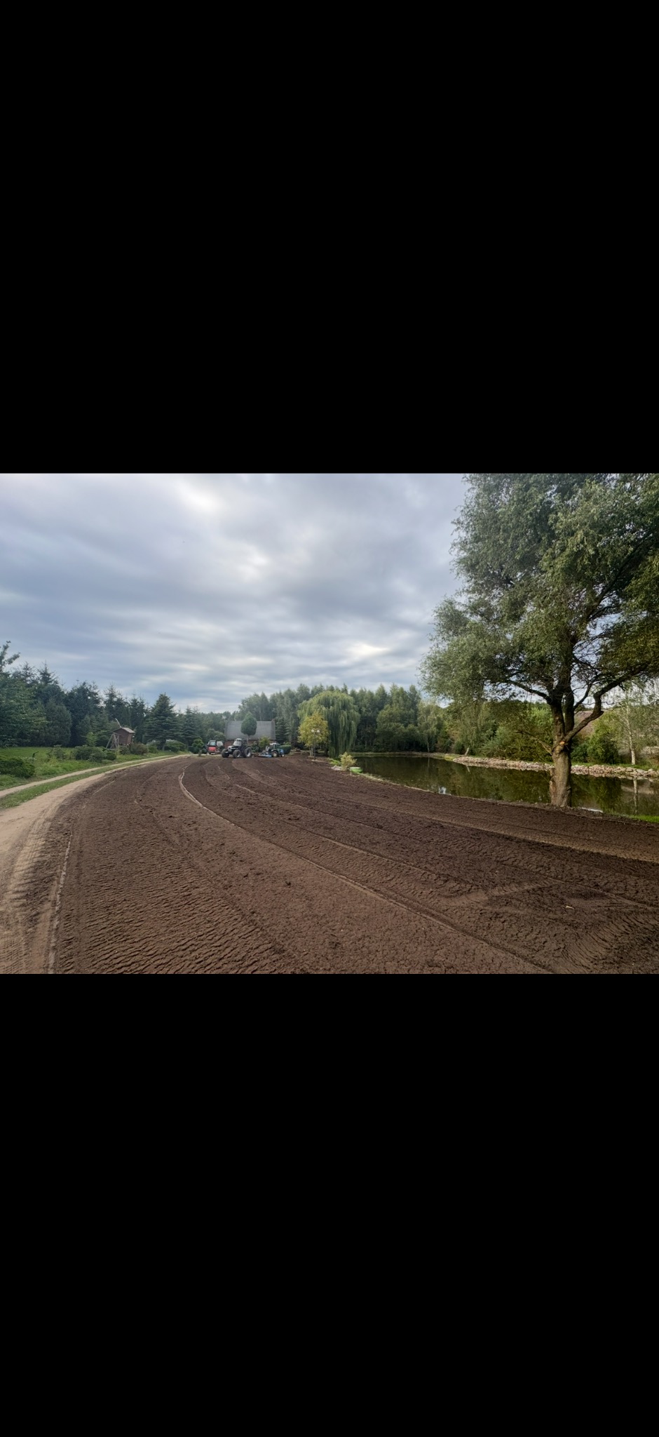 Przygotowany teren pod zasiew trawy, widoczne ślady opon, w tle maszyny rolnicze i mały staw. Pochmurne niebo.