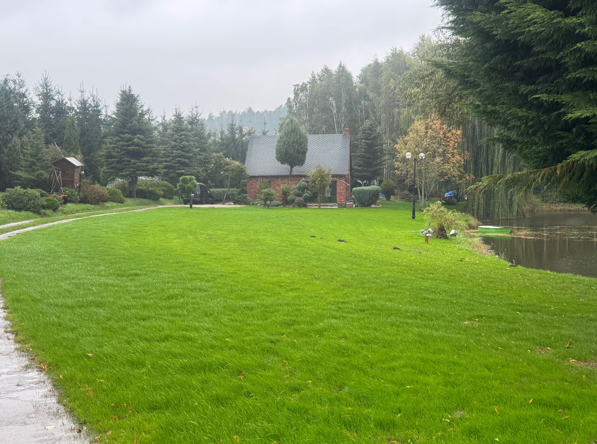 Zadbany trawnik z równo przystrzyżoną trawą, prowadzący do małego, ceglanego budynku. W tle drzewa i krzewy, a z boku fragment stawu. Widoczna ścieżka z kostki.