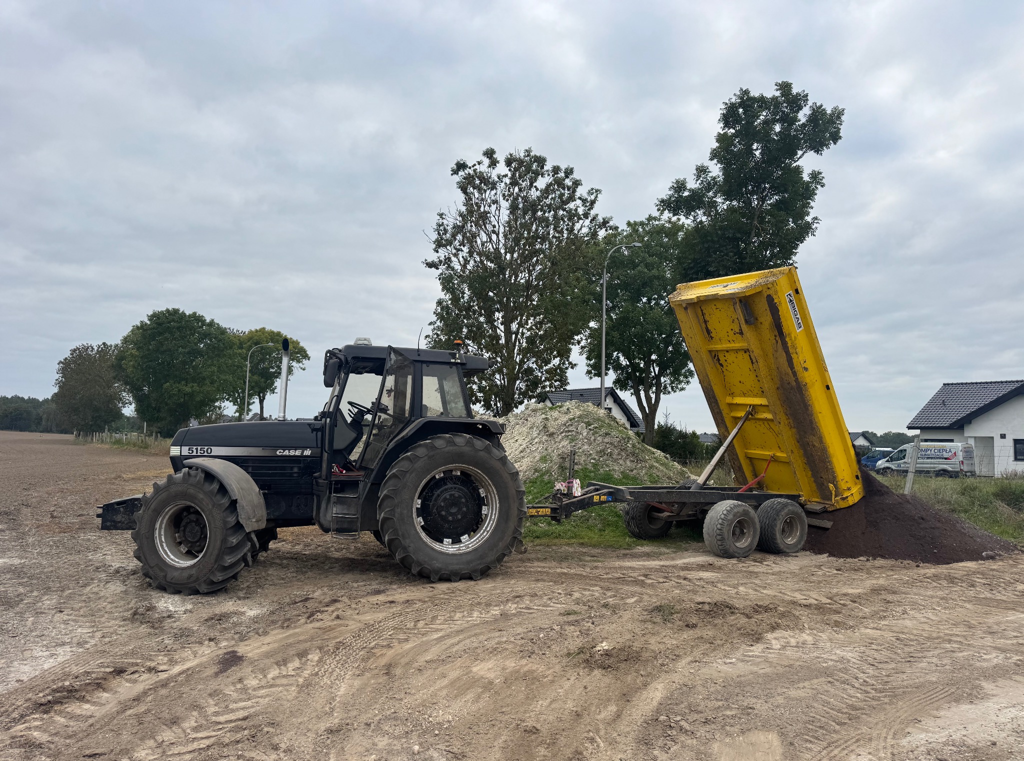Czarny traktor Case 5150 z żółtą przyczepą wywrotką, wysypujący ziemię na placu budowy. W tle drzewa i domy.