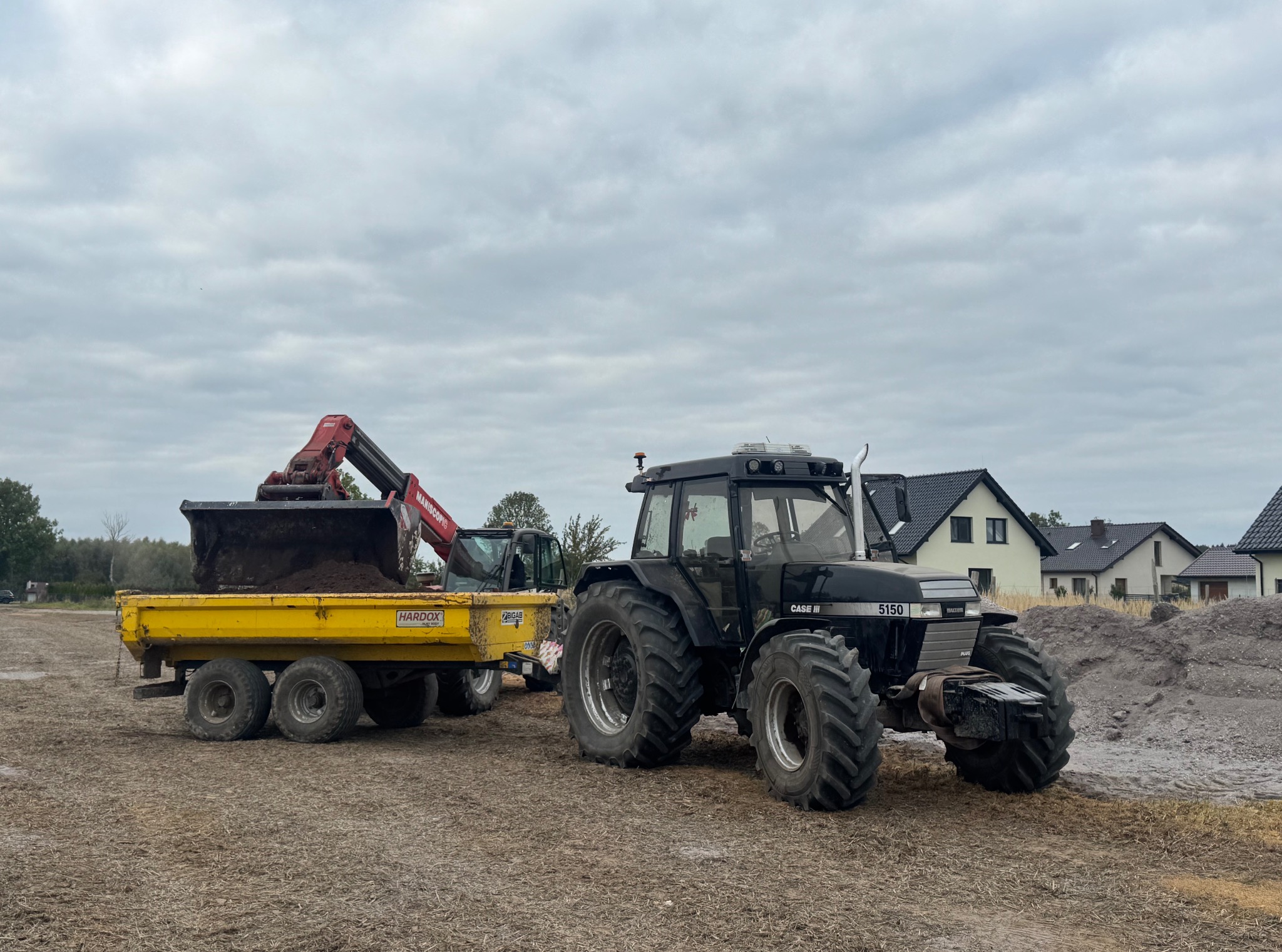 Czarny traktor Case 5150 z żółtą przyczepą, na którą koparka MANISCOPIC nabiera ziemię, w tle domy i pochmurne niebo. Prace ziemne w toku.
