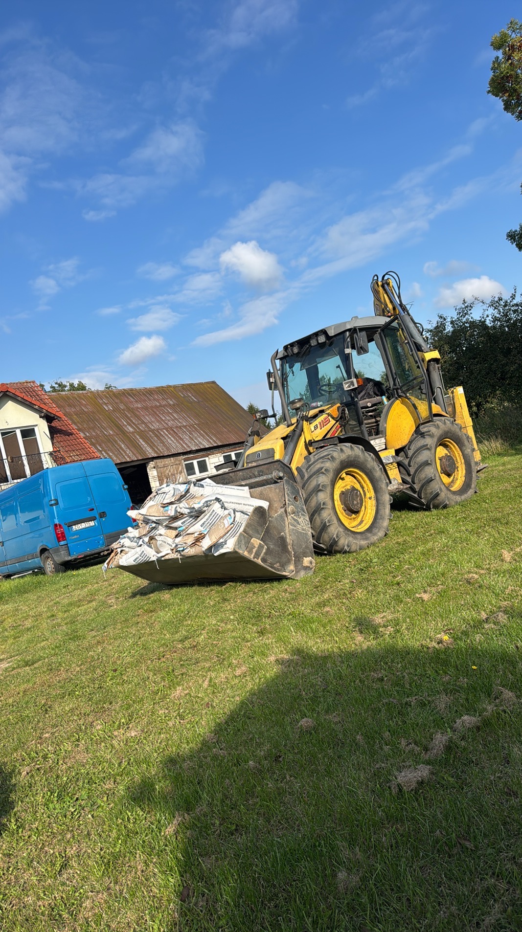Żółta koparko-ładowarka z pełną łyżką worków na trawiastym terenie, zaparkowany niebieski bus, budynek w tle, błękitne niebo z chmurami.