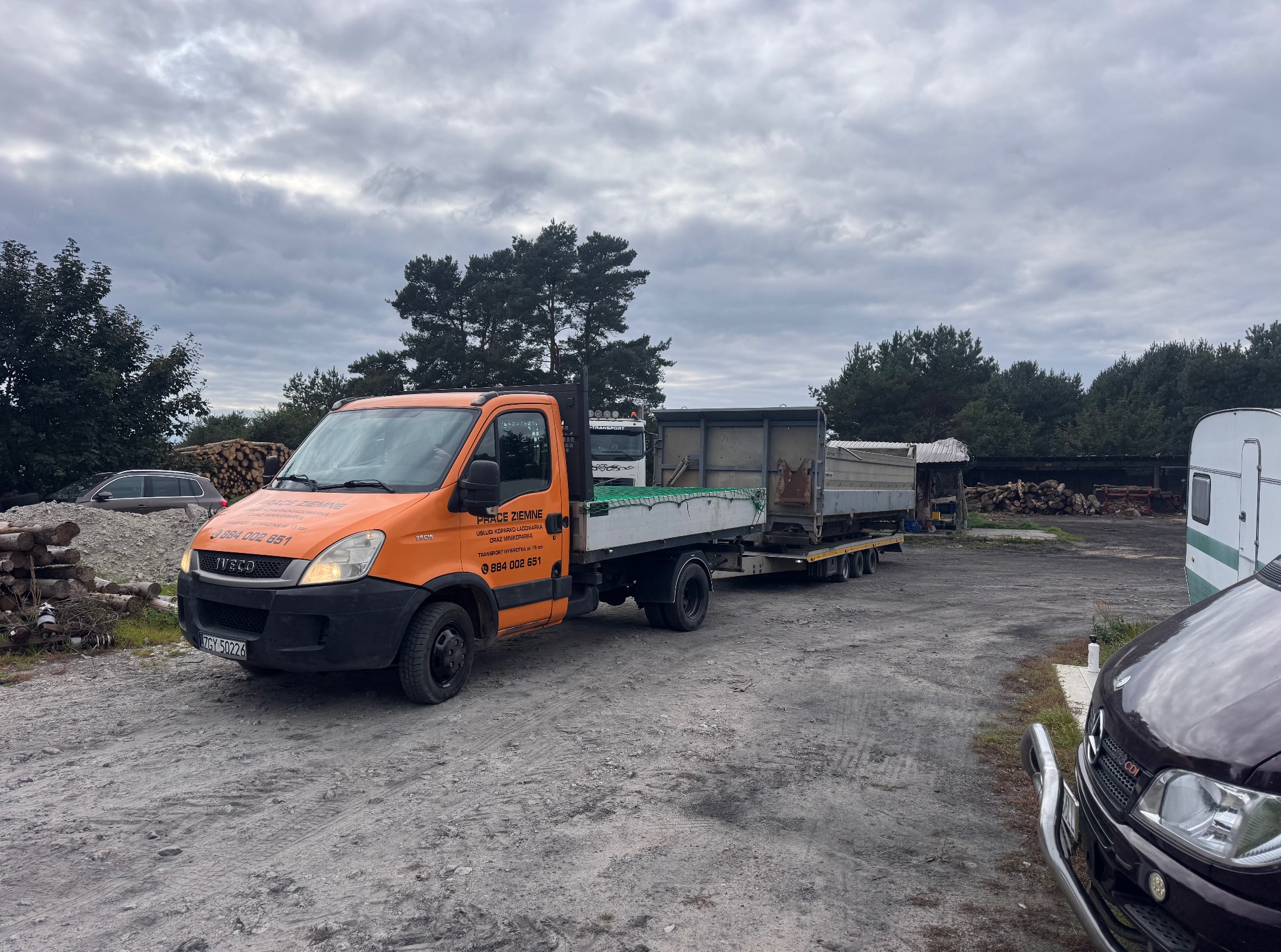 Pomarańczowy samochód ciężarowy z przyczepą i napisem 'Prace Ziemne' na tle stosu drewna i innych pojazdów na placu. Pochmurne niebo.