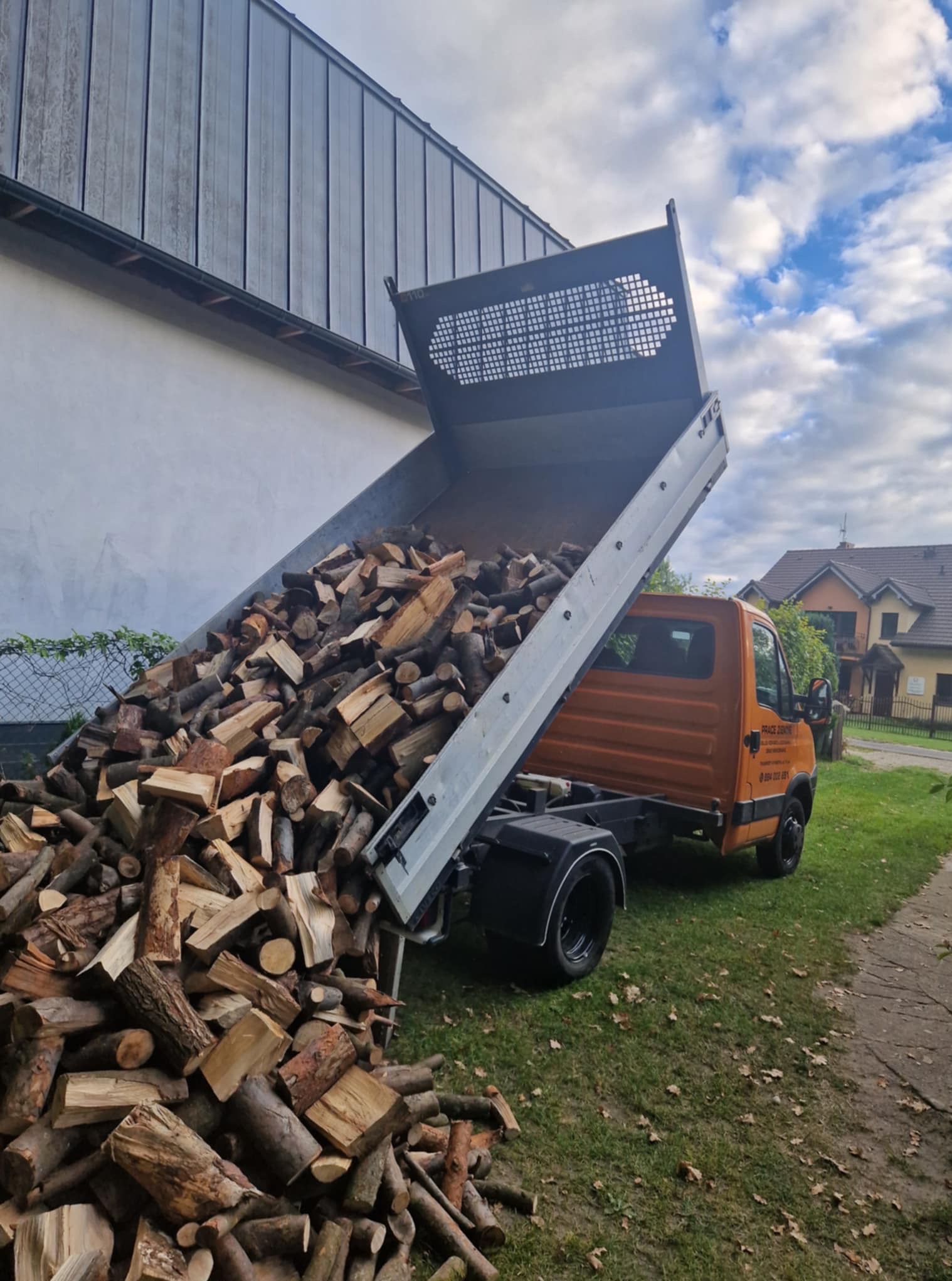 Pomarańczowa wywrotka z podniesioną skrzynią wyładowuje drewno w Gryficach. Widoczne pocięte kawałki drewna na trawie, budynek w tle i fragment nieba.