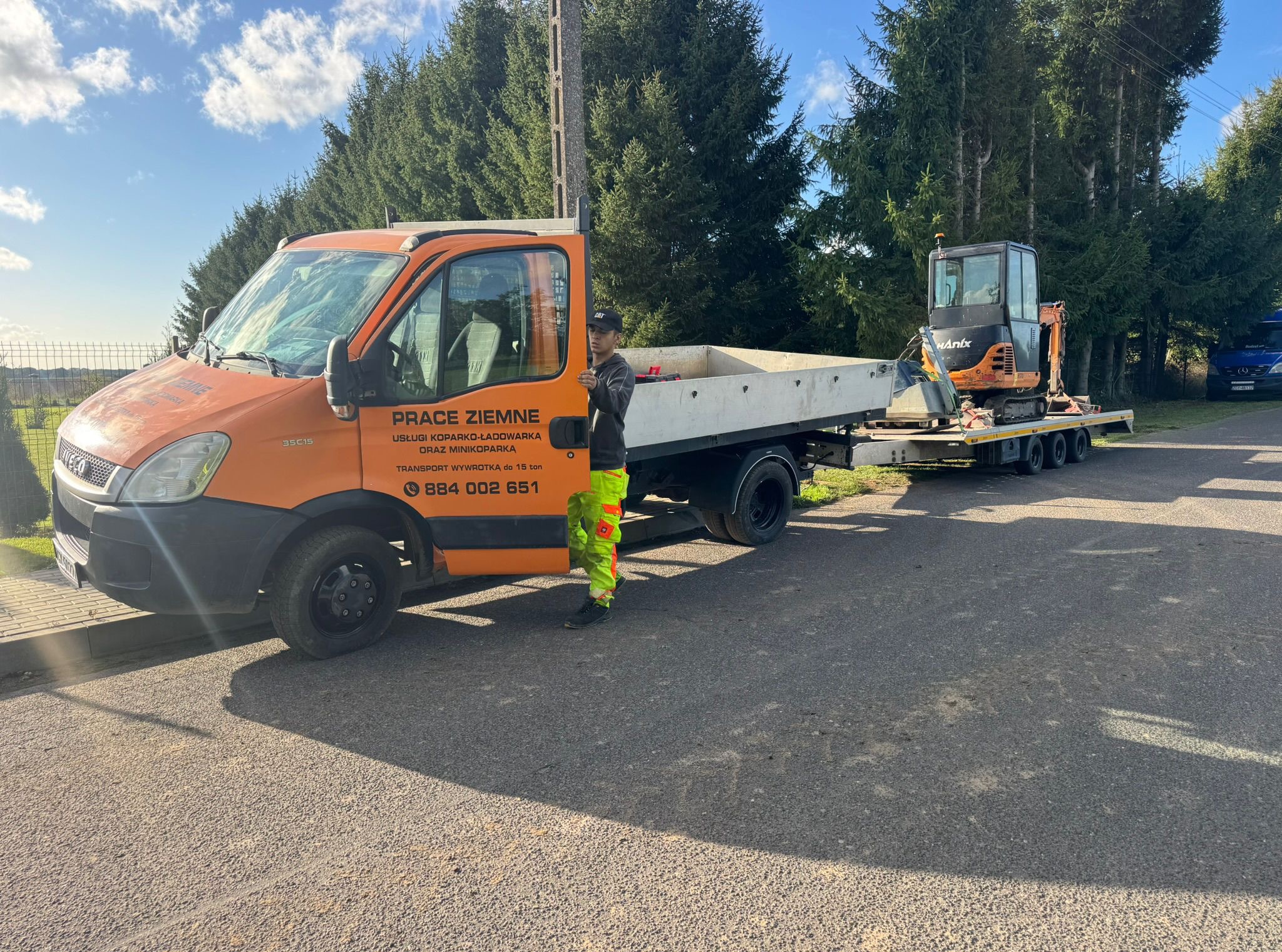 Pomarańczowa ciężarówka z przyczepą i małą koparką, pracownik w odblaskowym ubraniu na tle drzew. Napis na ciężarówce: Prace Ziemne, usługi koparko-ładowarką.