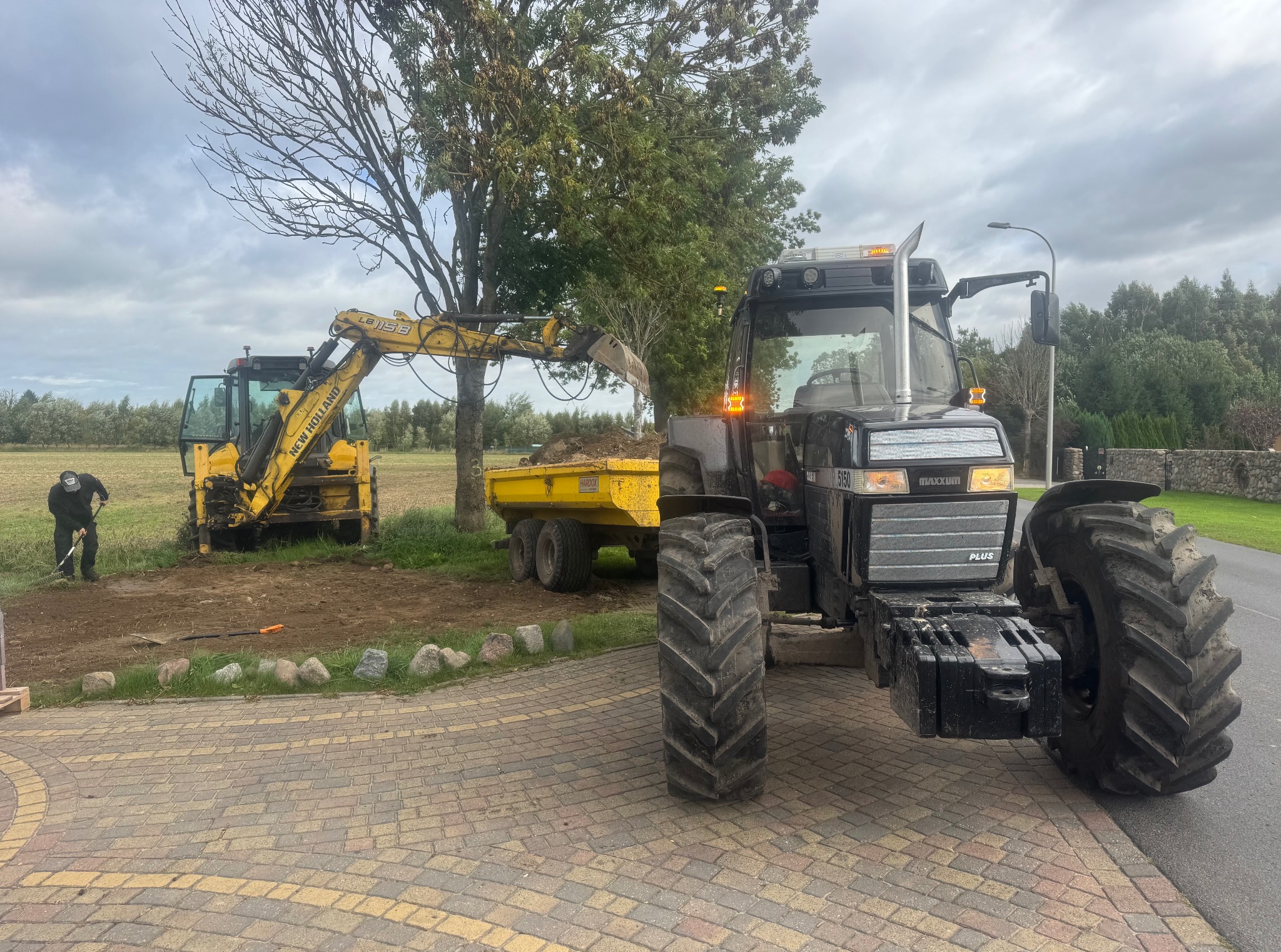 Żółta koparka ładuje ziemię na przyczepę ciągniętą przez traktor na tle drzew i pola. Pracownik z grabiami wyrównuje teren. Widok z kostki brukowej.