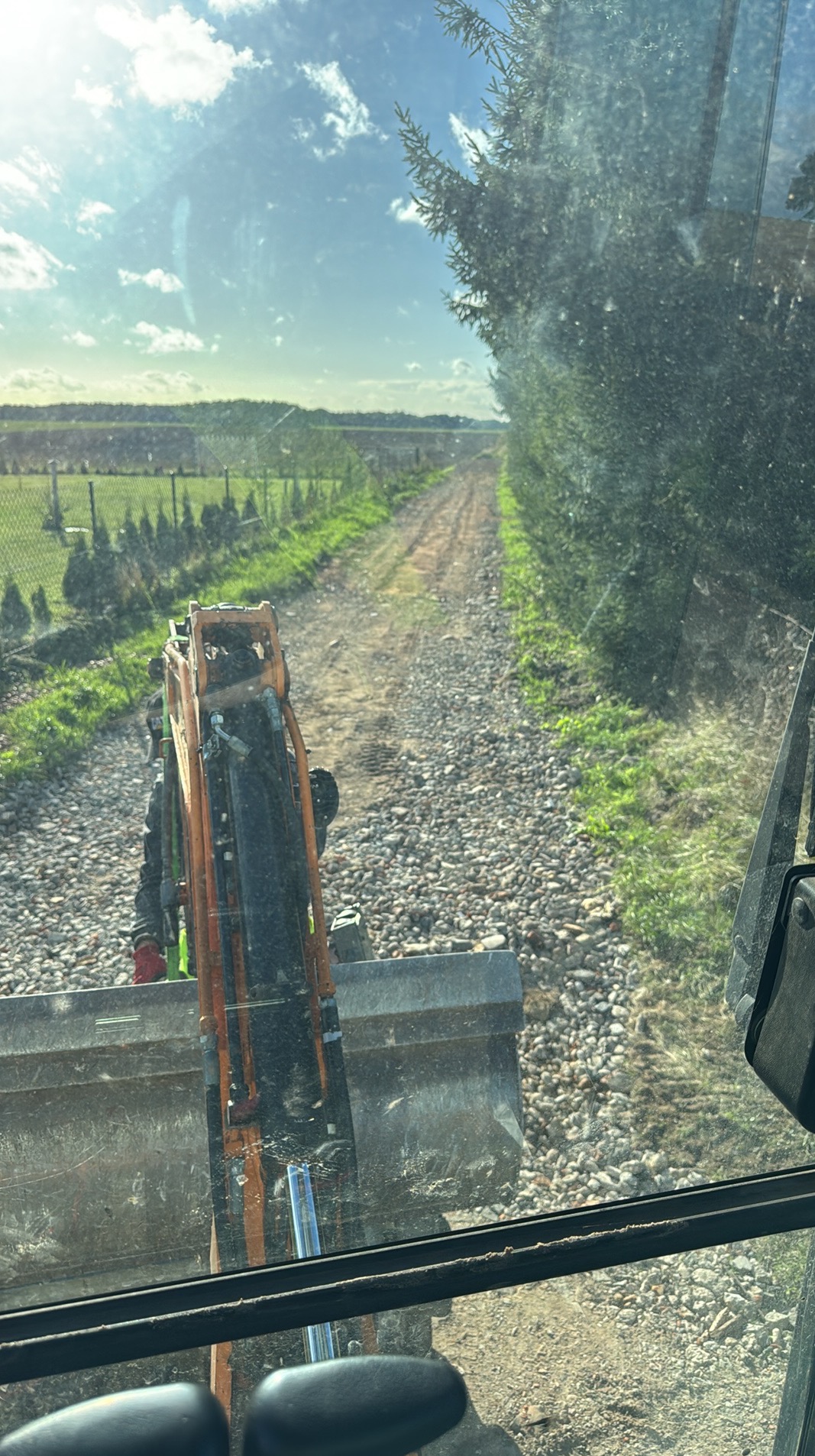 Widok z kabiny koparki na drogę z tłucznia, otoczoną zielenią i drzewami. W tle widoczne pole i niebo. Perspektywa z wnętrza maszyny.