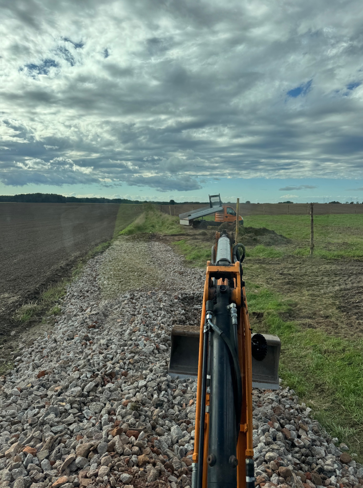 Widok z kabiny koparki na utwardzoną żwirem drogę polną, z pomarańczową ciężarówką w tle, pod pochmurnym niebem. Perspektywa operatora maszyny.