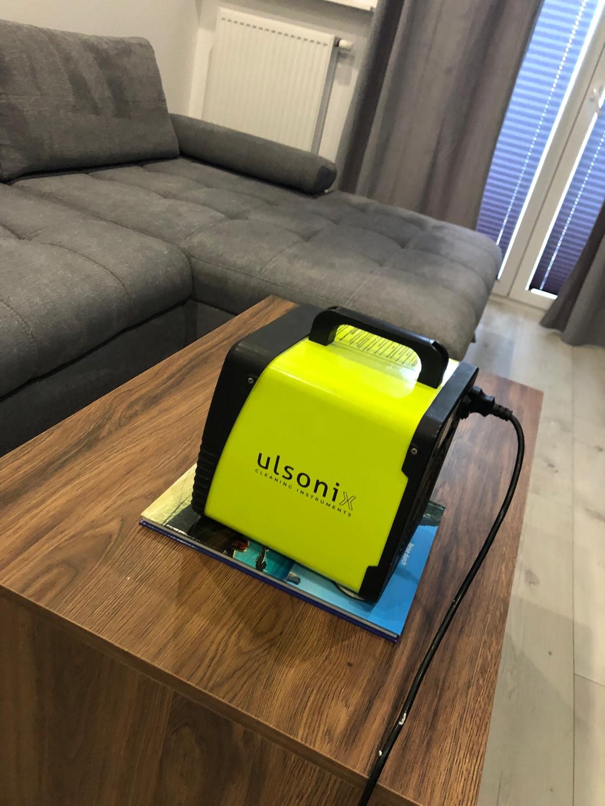 Żółty sprzęt czyszczący Ulsonix na drewnianym stoliku, obok szarej sofy i okna z żaluzjami. Kabel zasilający widoczny. Sprzęt do czyszczenia tapicerki.
