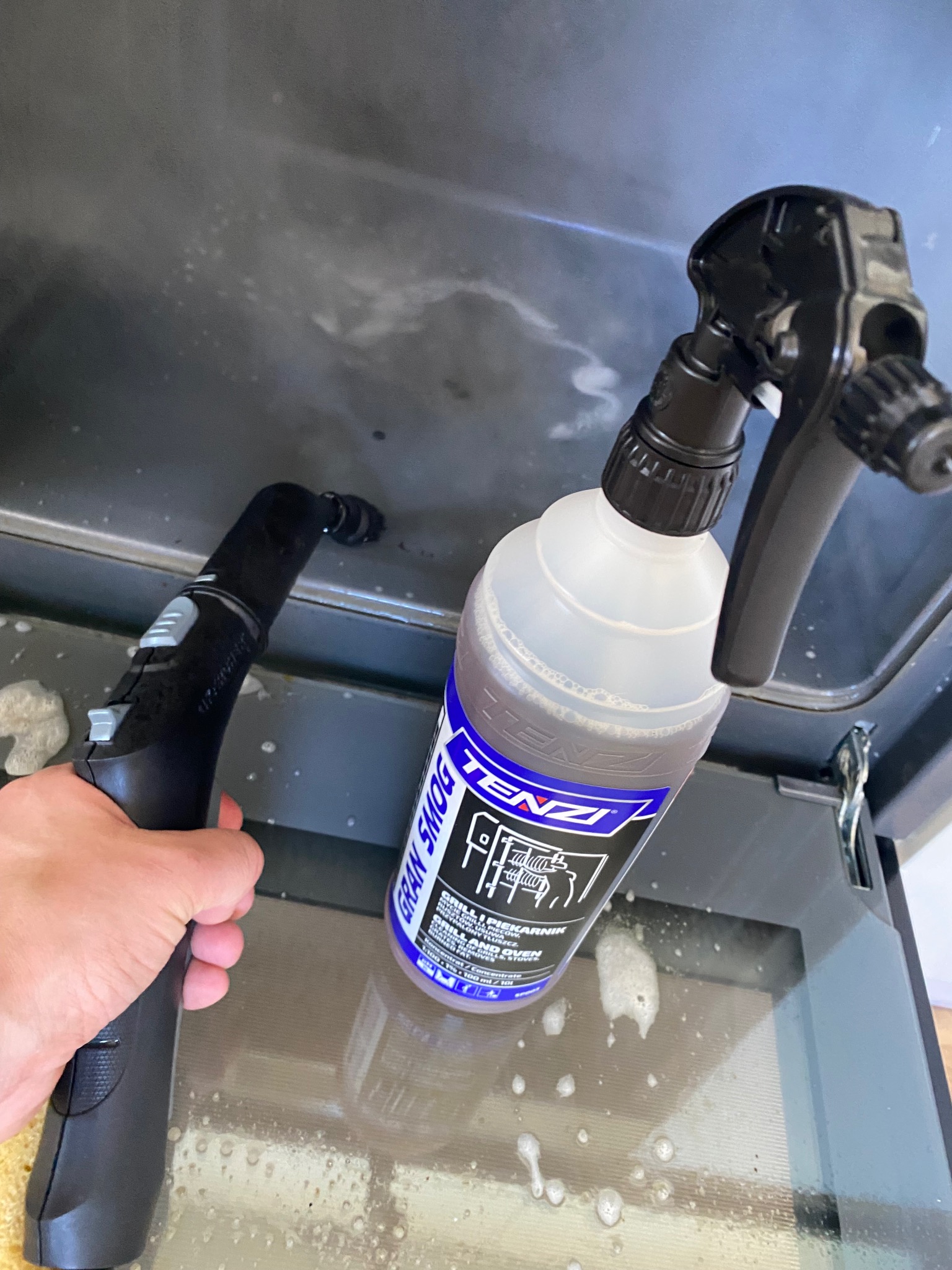 Czyszczenie piekarnika parownicą i detergentem Tenzi Gran Smog, widoczne krople płynu na szklanych drzwiach piekarnika podczas usuwania zabrudzeń.