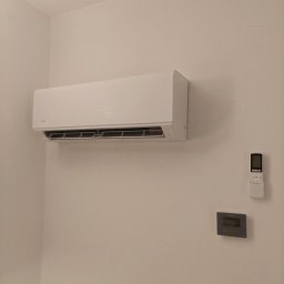 SOFTWARE PIZZA SPÓŁKA Z OGRANICZONĄ ODPOWIEDZIALNOŚCIĄ - Biały klimatyzator ścienny marki AUX, zamontowany w pomieszczeniu. Obok widoczny pilot i panel sterowania. Minimalistyczny design.