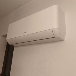 SOFTWARE PIZZA SPÓŁKA Z OGRANICZONĄ ODPOWIEDZIALNOŚCIĄ - Biała jednostka klimatyzacyjna Kaisai zamontowana na jasnej ścianie w pomieszczeniu. Widoczny fragment drzwi po lewej stronie kadru. Minimalistyczny design.