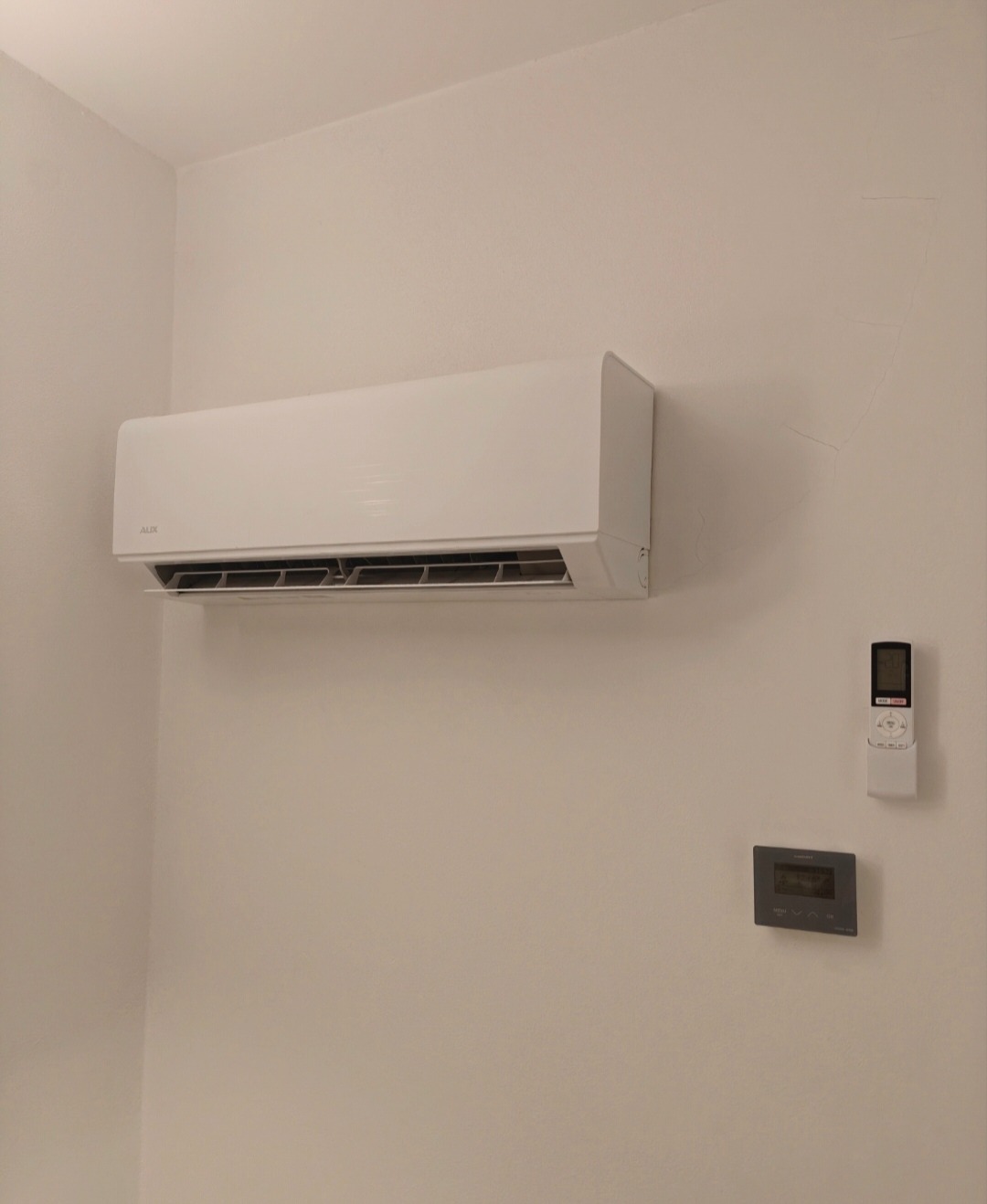 Biały klimatyzator ścienny marki AUX, zamontowany w pomieszczeniu. Obok widoczny pilot i panel sterowania. Minimalistyczny design.