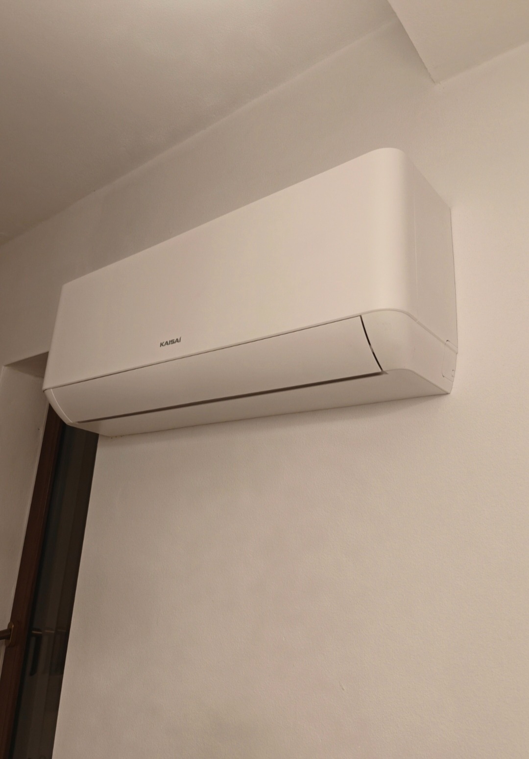Biała jednostka klimatyzacyjna Kaisai zamontowana na jasnej ścianie w pomieszczeniu. Widoczny fragment drzwi po lewej stronie kadru. Minimalistyczny design.
