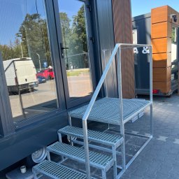 Remigiusz Ducki - Ocynkowane metalowe schody kratownicowe z podestem i barierką, prowadzące do wejścia ciemnoszarego kontenera biurowego z dużymi szklanymi drzwiami. W tle drugi kontener z drewnopodobną okładziną.