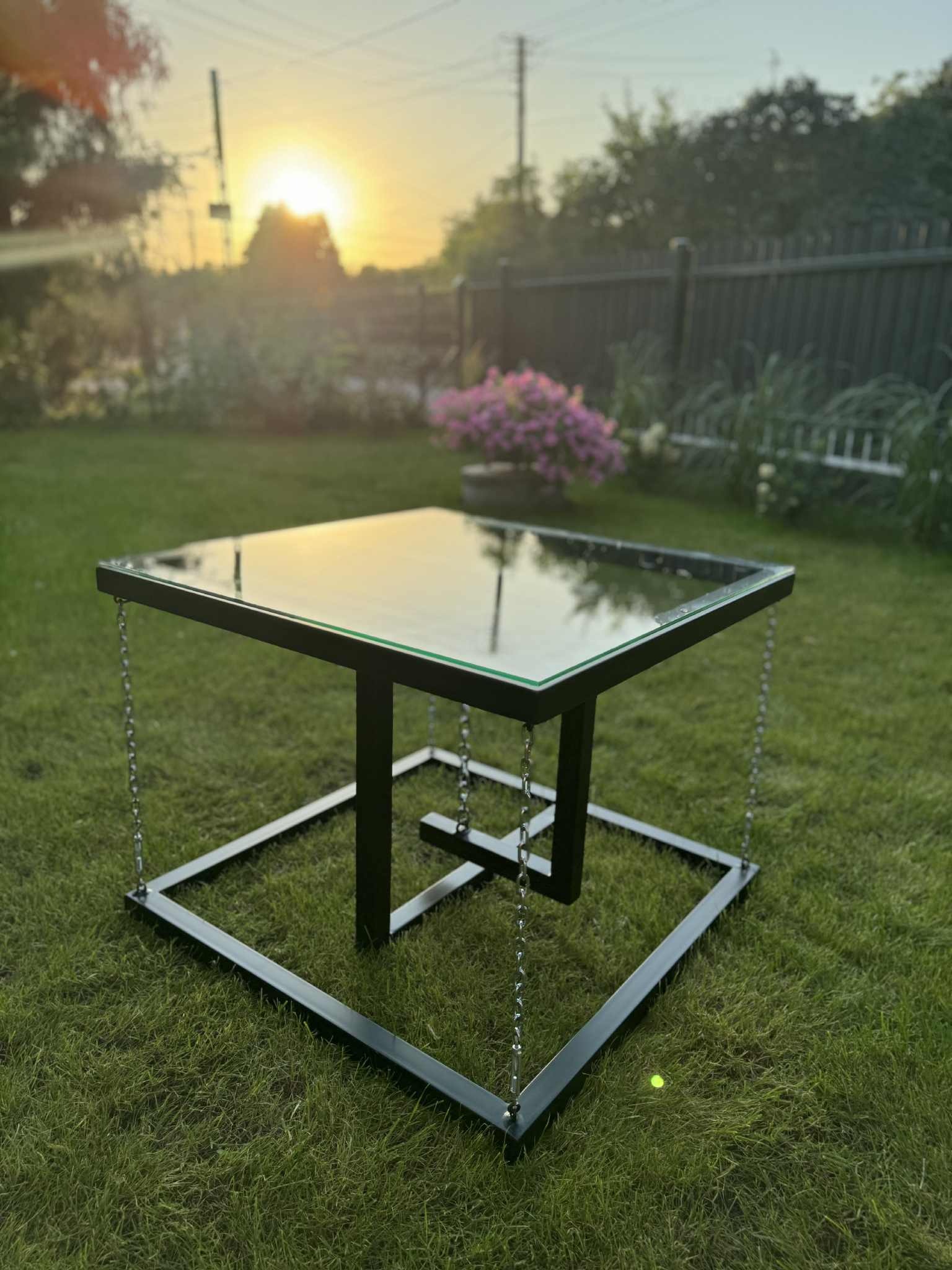 Nietypowy stolik kawowy z metalową konstrukcją i szklanym blatem, zawieszony na łańcuchach, na tle zielonego trawnika w promieniach zachodzącego słońca.