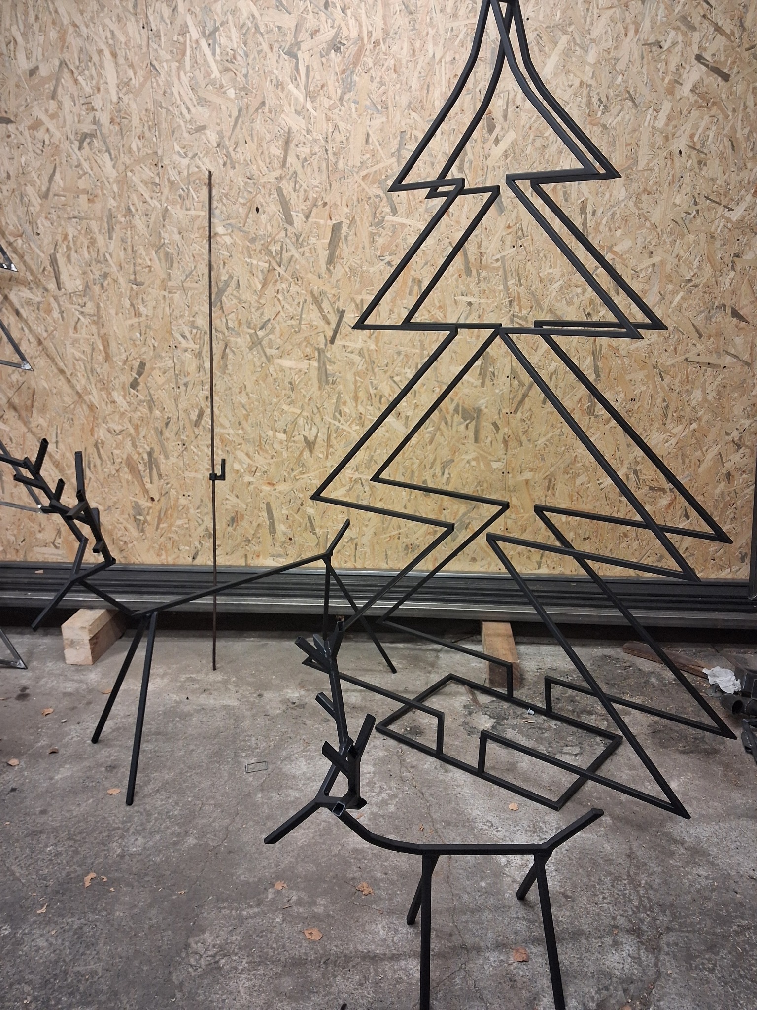 Metalowe konstrukcje: choinka i renifery z czarnych prętów na tle płyty OSB. Elementy dekoracyjne w surowym warsztatowym otoczeniu, gotowe do dalszej obróbki.