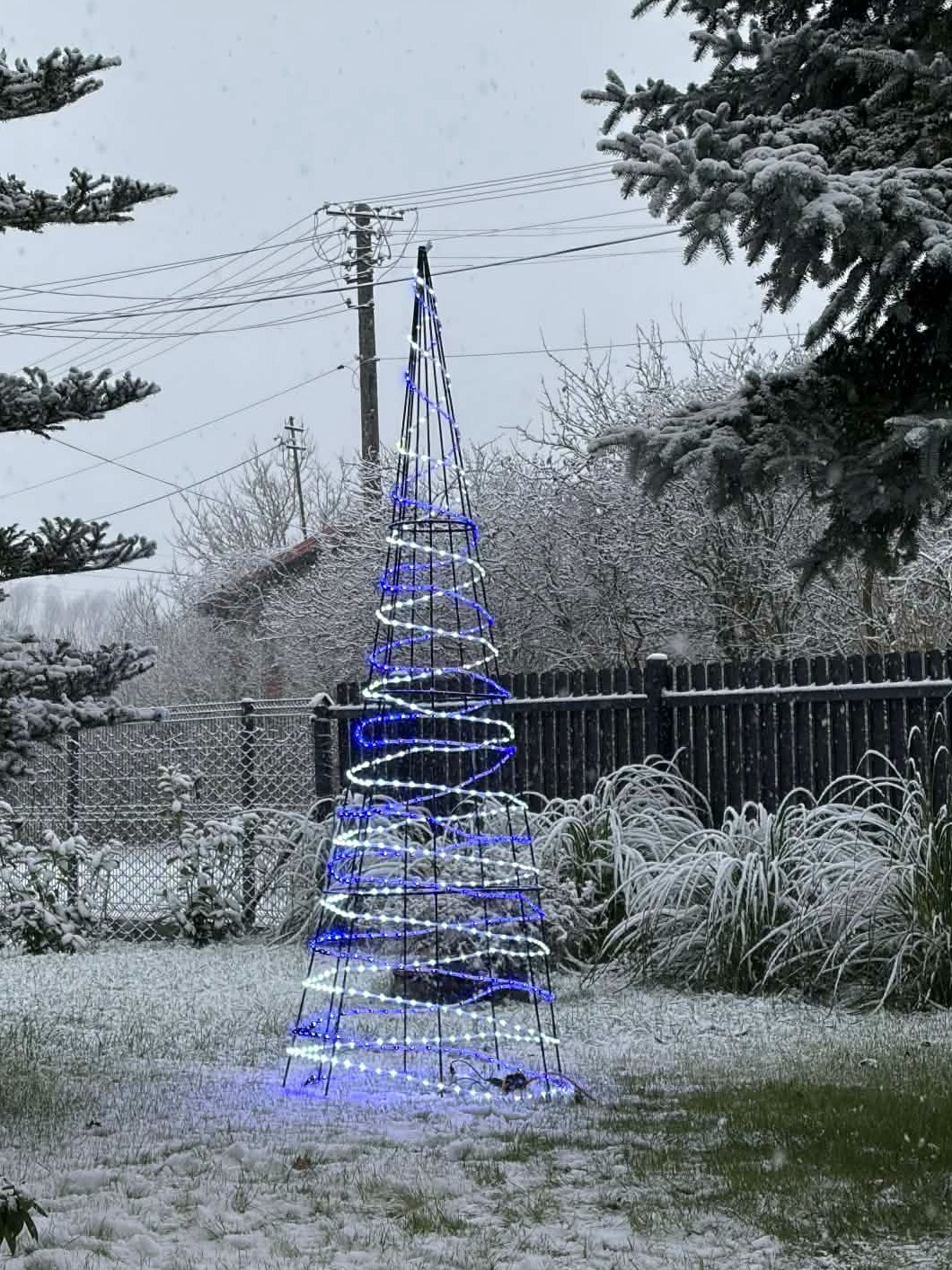 Metalowa konstrukcja choinki z niebiesko-białymi światełkami LED, stojąca na ośnieżonym trawniku w zimowej scenerii. W tle płot i drzewa pokryte śniegiem.