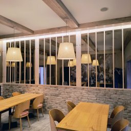 Los Deskos - Jasne drewniane belki sufitowe i ażurowa ścianka działowa z drewna w restauracji. Widoczne stoły i krzesła, lampy z abażurami. Ścianka z cegły. W tle osoba robiąca zdjęcie.