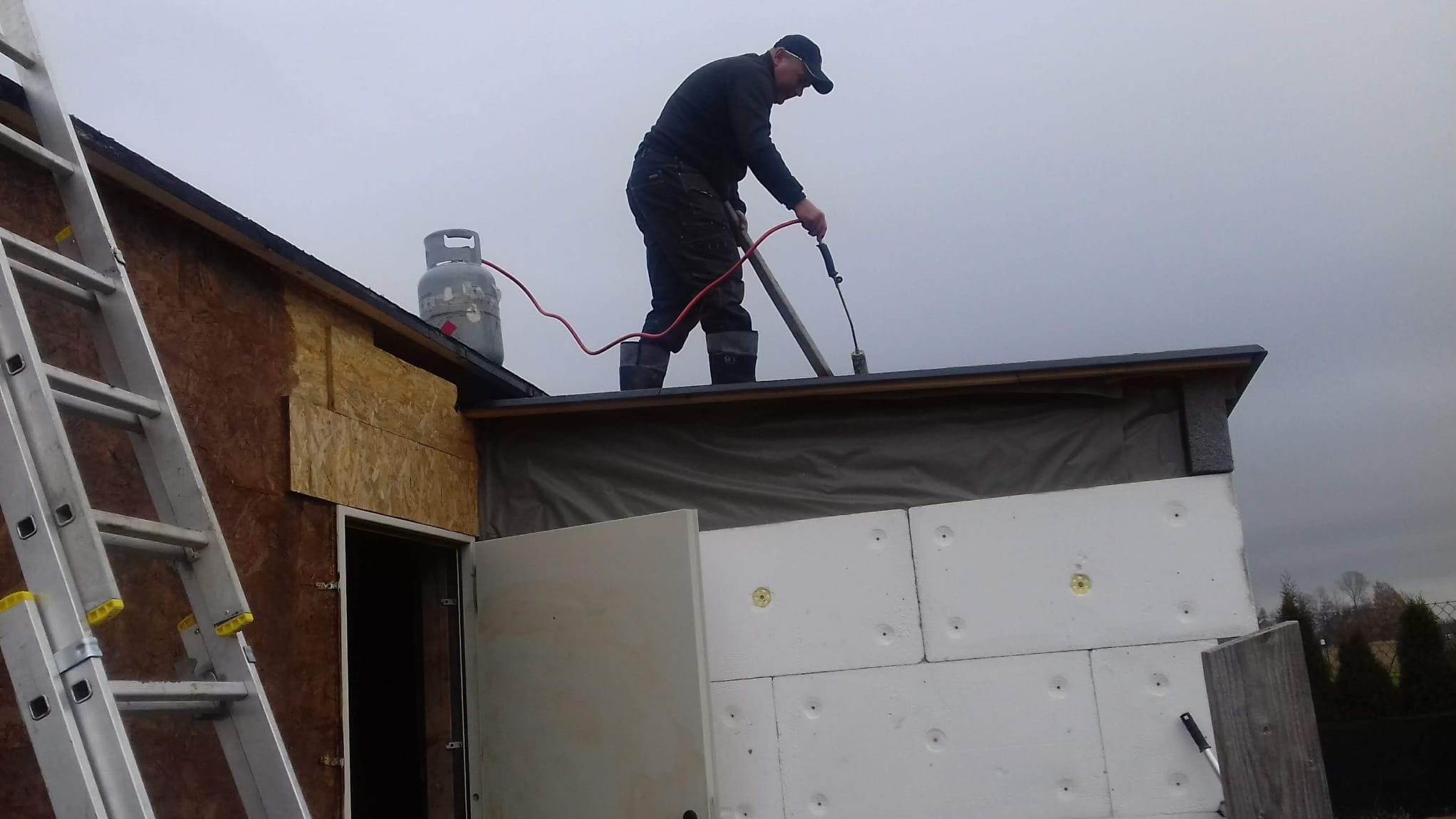 Pracownik w czarnym ubraniu na dachu budynku ociepla go styropianem. Widoczna butla gazowa, drabina i narzędzia. Praca na wysokościach.