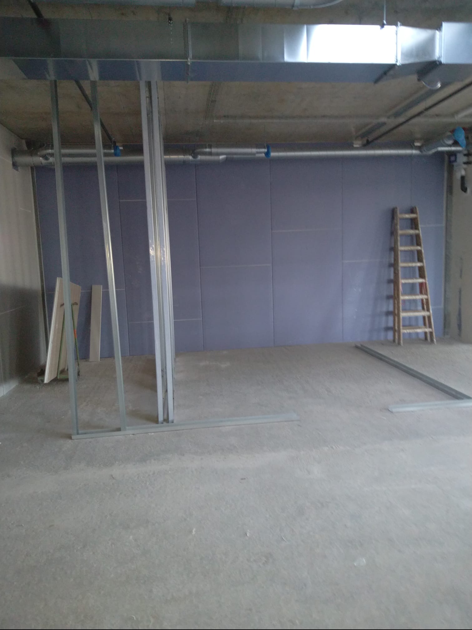 Montaż ścian z płyt karton-gips w surowym wnętrzu. Widoczne metalowe profile, płyty GK na ścianie, drabina i instalacje pod sufitem. Etap prac wykończeniowych.