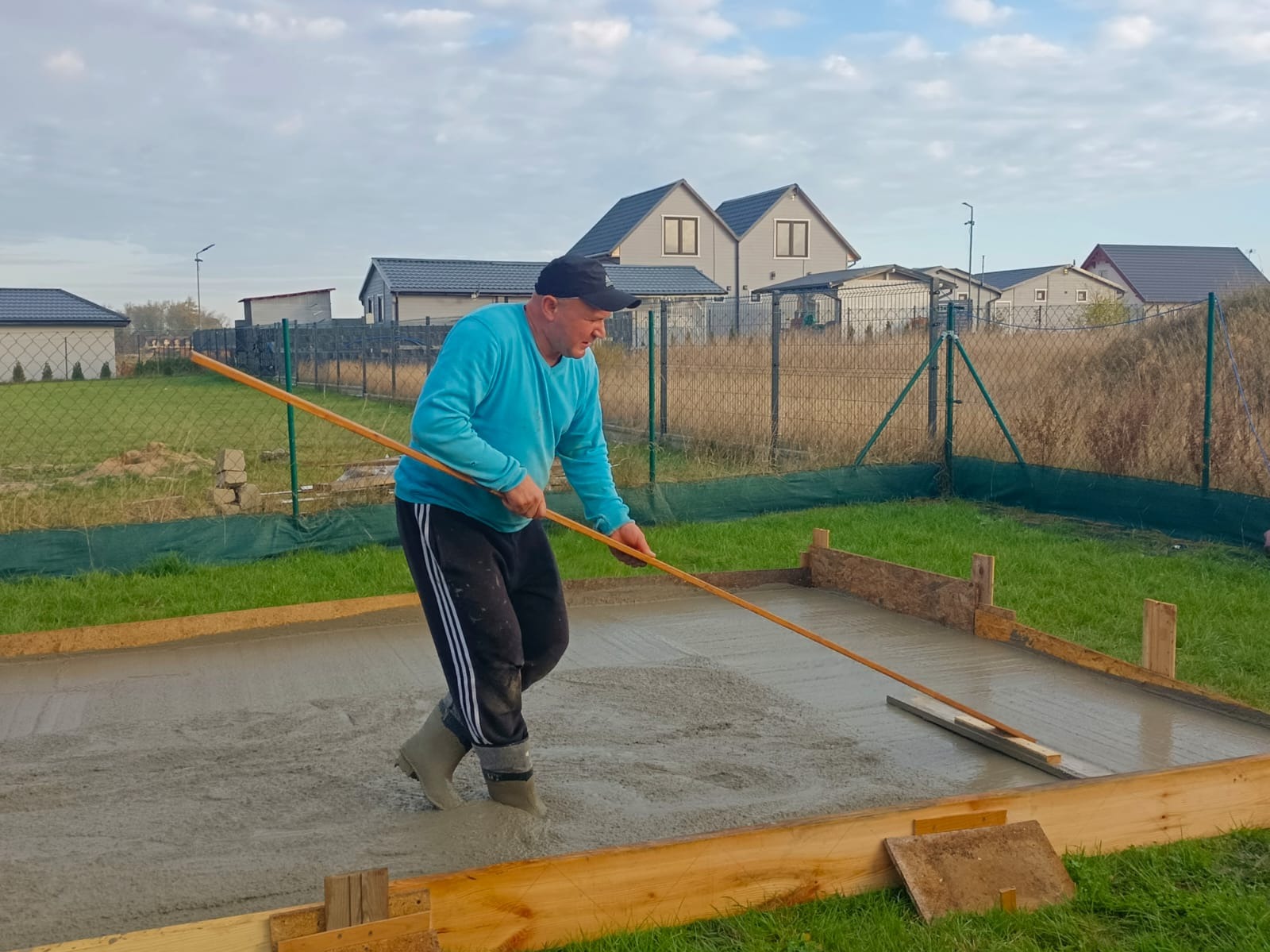 Pracownik w niebieskiej bluzie wyrównuje świeżo wylaną, szarą posadzkę w drewnianym szalunku na zewnątrz, w tle domy jednorodzinne i zielone trawniki.