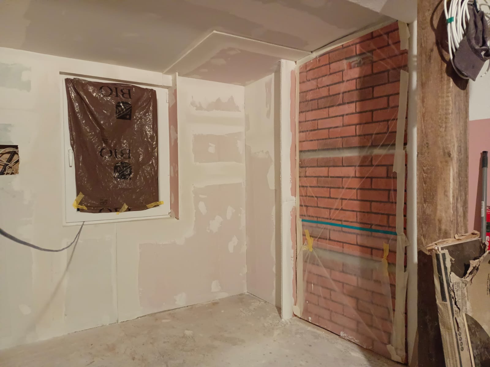 Pokój w trakcie prac wykończeniowych: ściany po szpachlowaniu, okno zabezpieczone folią, imitacja cegły na ścianie osłonięta folią malarską.