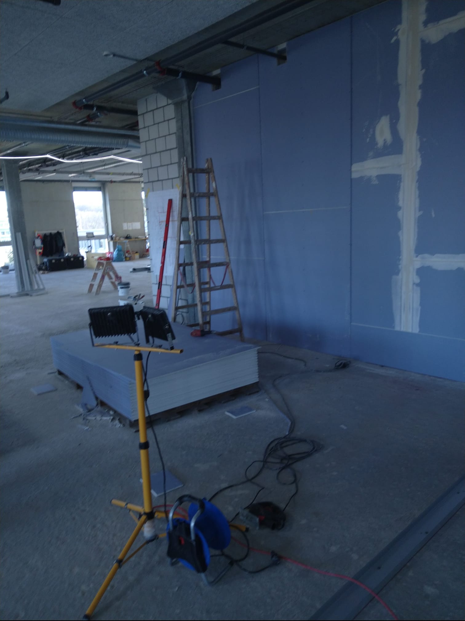 Niebieska ściana z płyt kartonowo-gipsowych w trakcie szpachlowania, drabina oparta o ścianę, lampa budowlana na pierwszym planie, widok na surowy stan wykończenia.