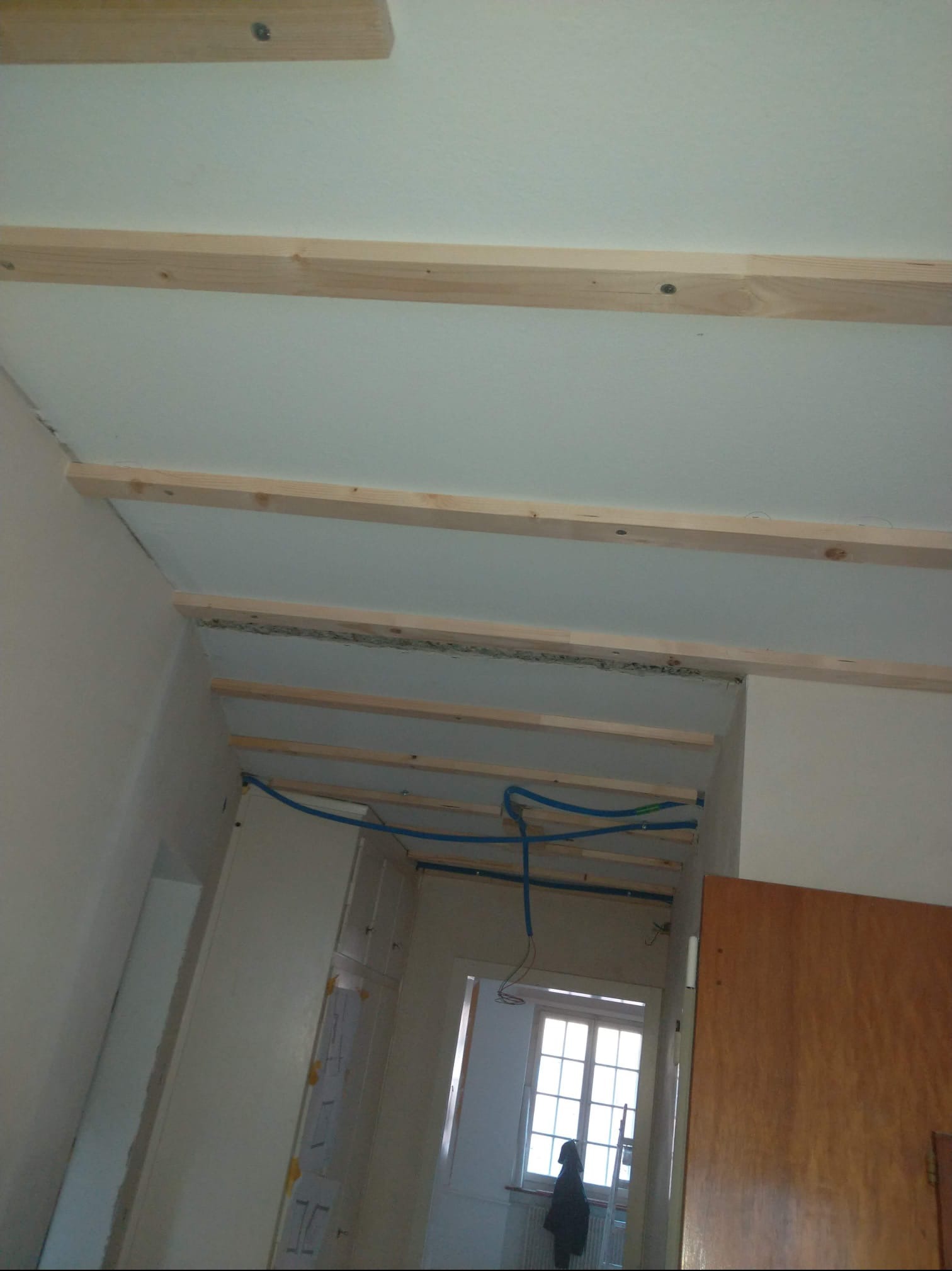 Sufit podwieszany z drewnianą konstrukcją i instalacją wodną w trakcie montażu. Widoczne elementy wykończeniowe i okno w tle. Ujęcie z dołu.