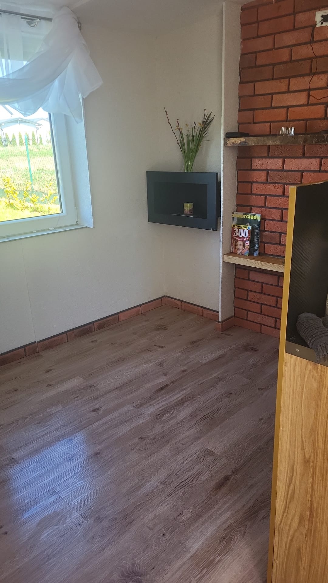 Jasne panele podłogowe w pokoju z ceglanym wykończeniem ściany i oknem z firanką. Na ścianie półka z dekoracjami i czasopismami.