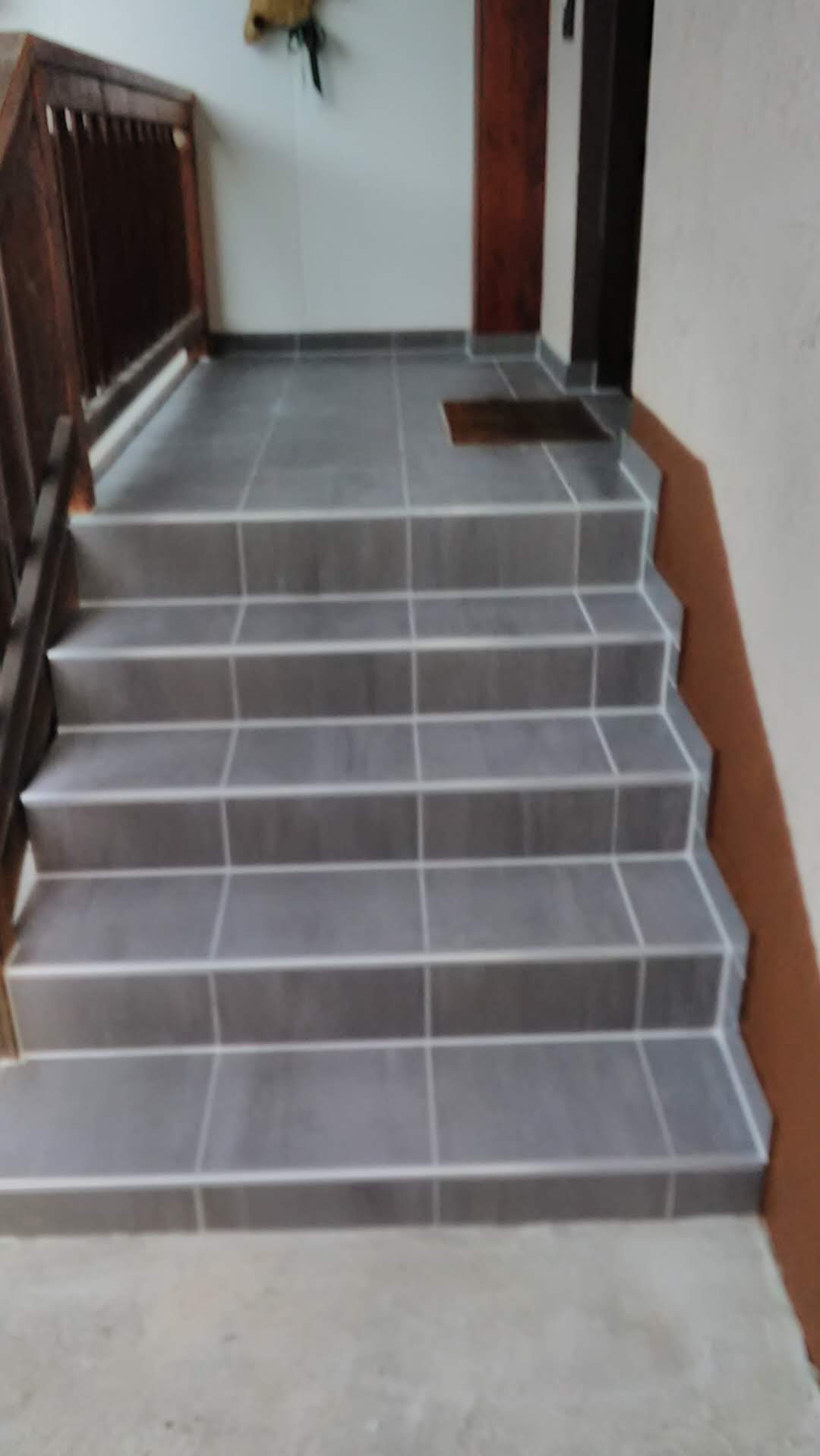 Schody wyłożone szarymi płytkami z jasną fugą, widok z dołu. Balustrada z ciemnego drewna po lewej stronie. Wejście do budynku.