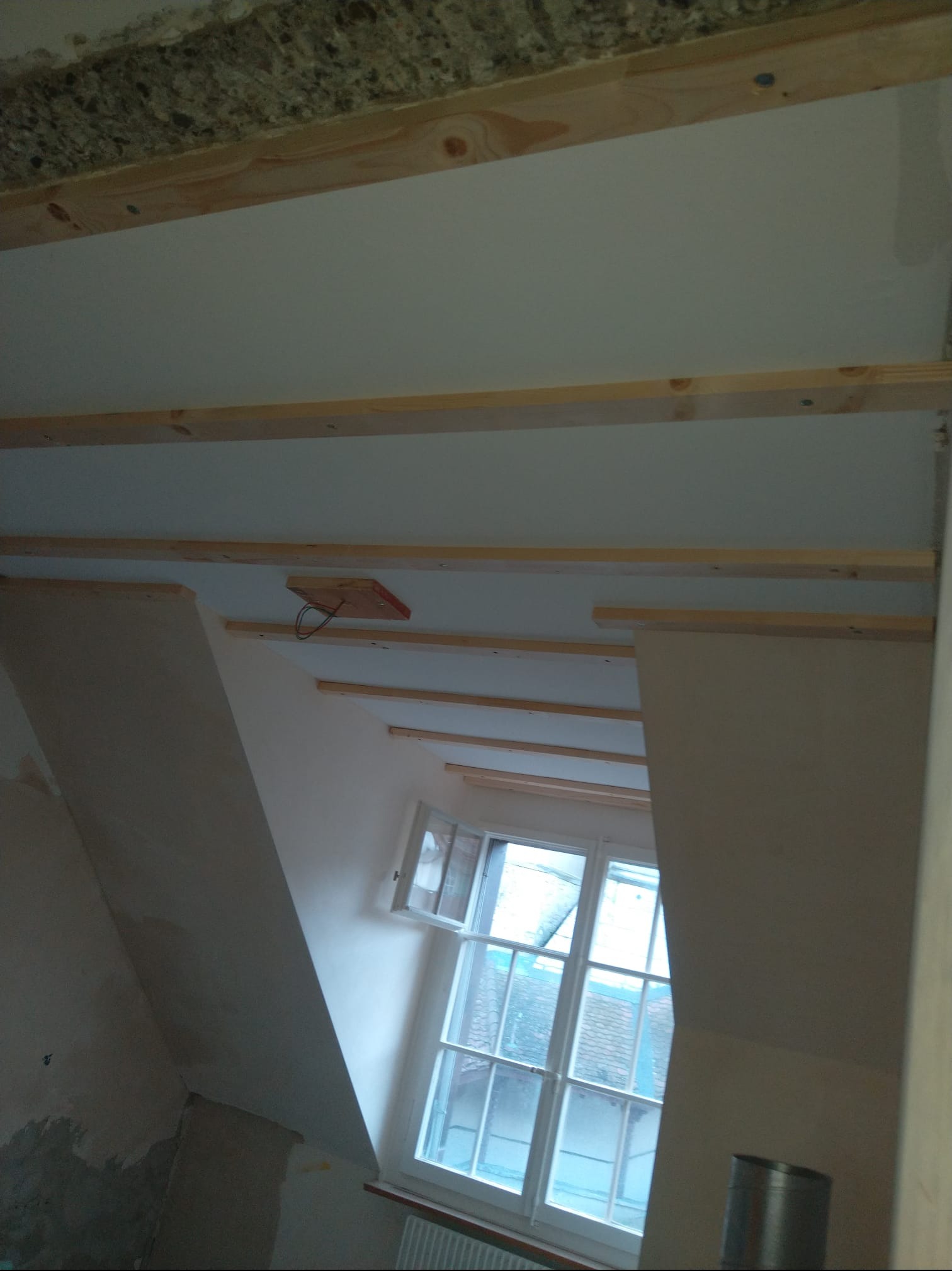 Skośny pokój na poddaszu w trakcie prac wykończeniowych. Widoczne drewniane belki, płyty gipsowe i okno. Fragment instalacji elektrycznej.
