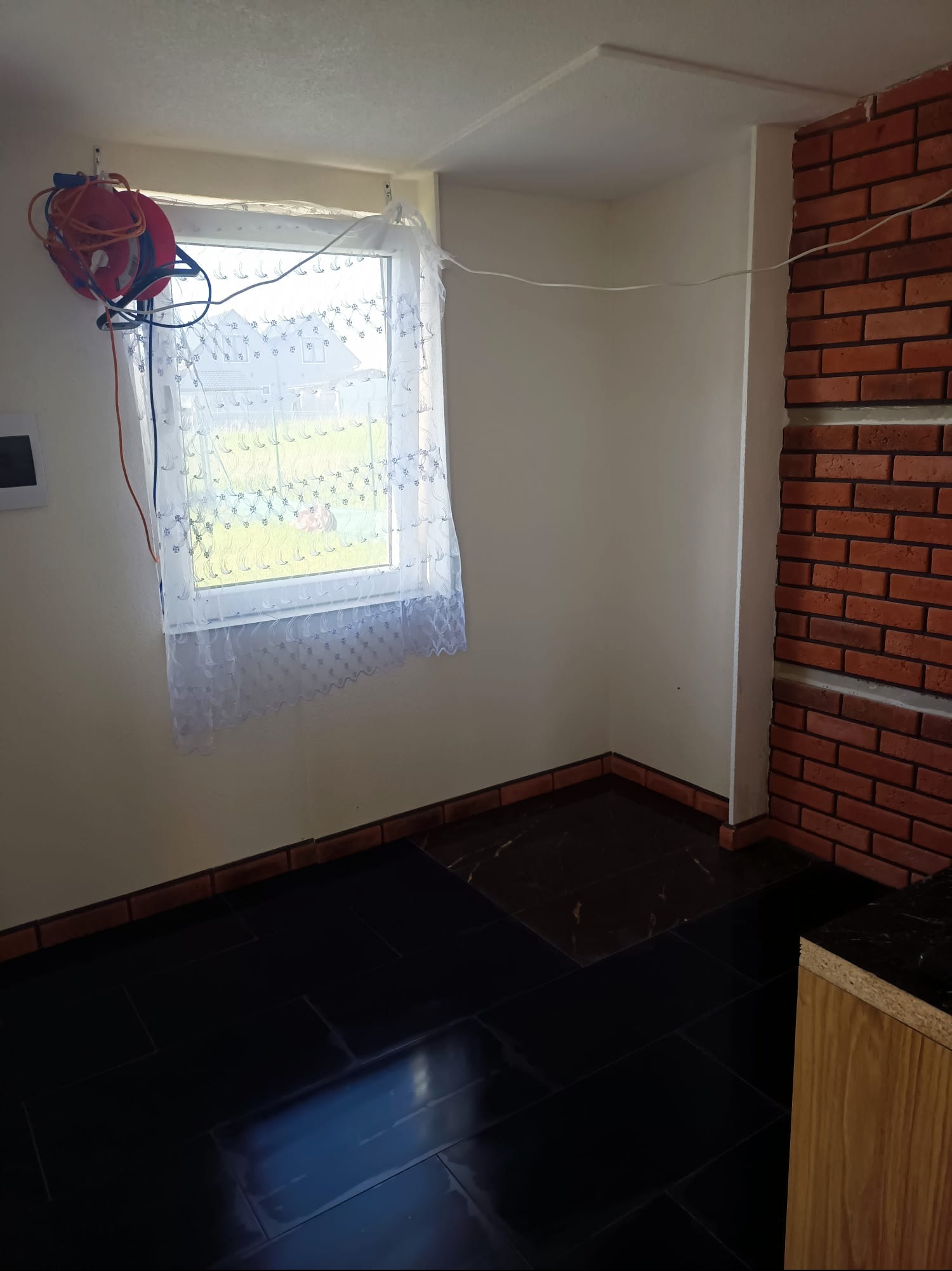 Wykończone wnętrze pokoju z czarną, błyszczącą podłogą z płytek, ceglaną ścianą i oknem z firanką. Widoczny zwinięty przedłużacz na ścianie.