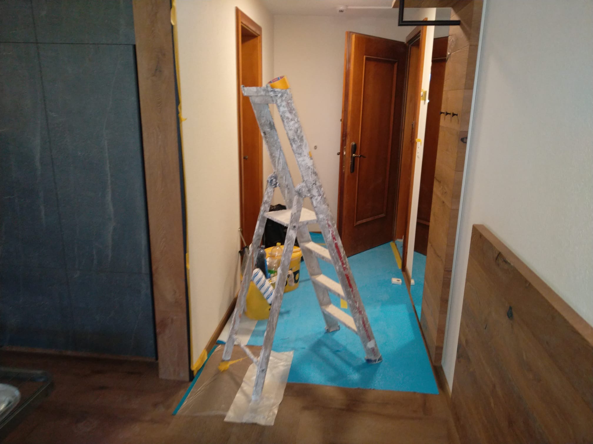 Wnętrze w trakcie prac: drabina malarska, zabezpieczona podłoga folią i taśmą. Widoczne otwarte drzwi i fragmenty mebli. Przygotowanie do malowania ścian.