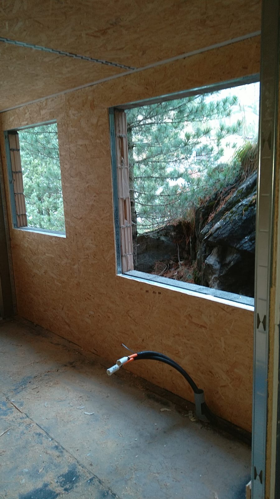 Wnętrze w trakcie budowy z płyt OSB, widoczne okna z widokiem na zieleń i skały, oraz instalacja elektryczna w podłodze. Metalowe profile konstrukcyjne.