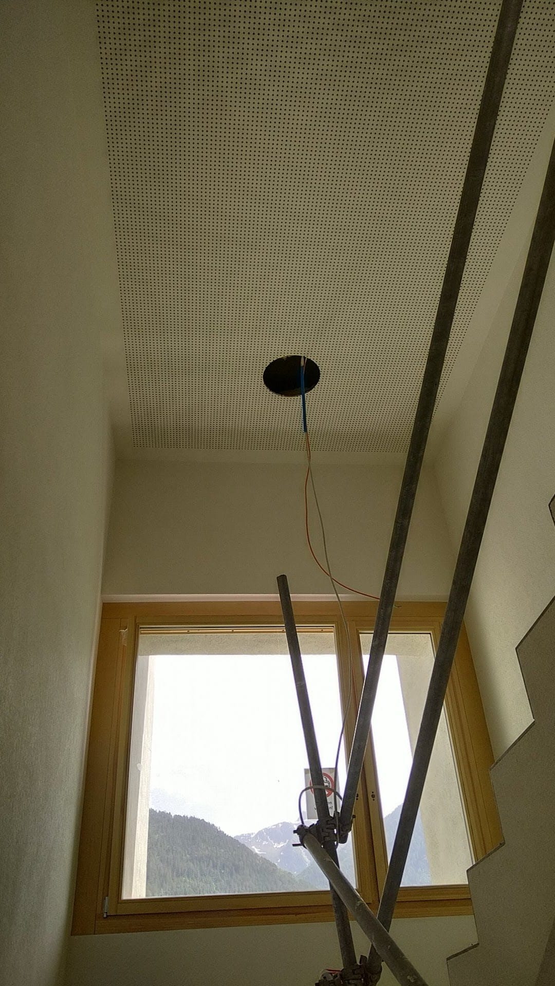 Sufit podwieszany z perforacją i otworem na oświetlenie, widok z dołu, fragment rusztowania, okno z widokiem na góry w tle. Prace wykończeniowe w pomieszczeniu.