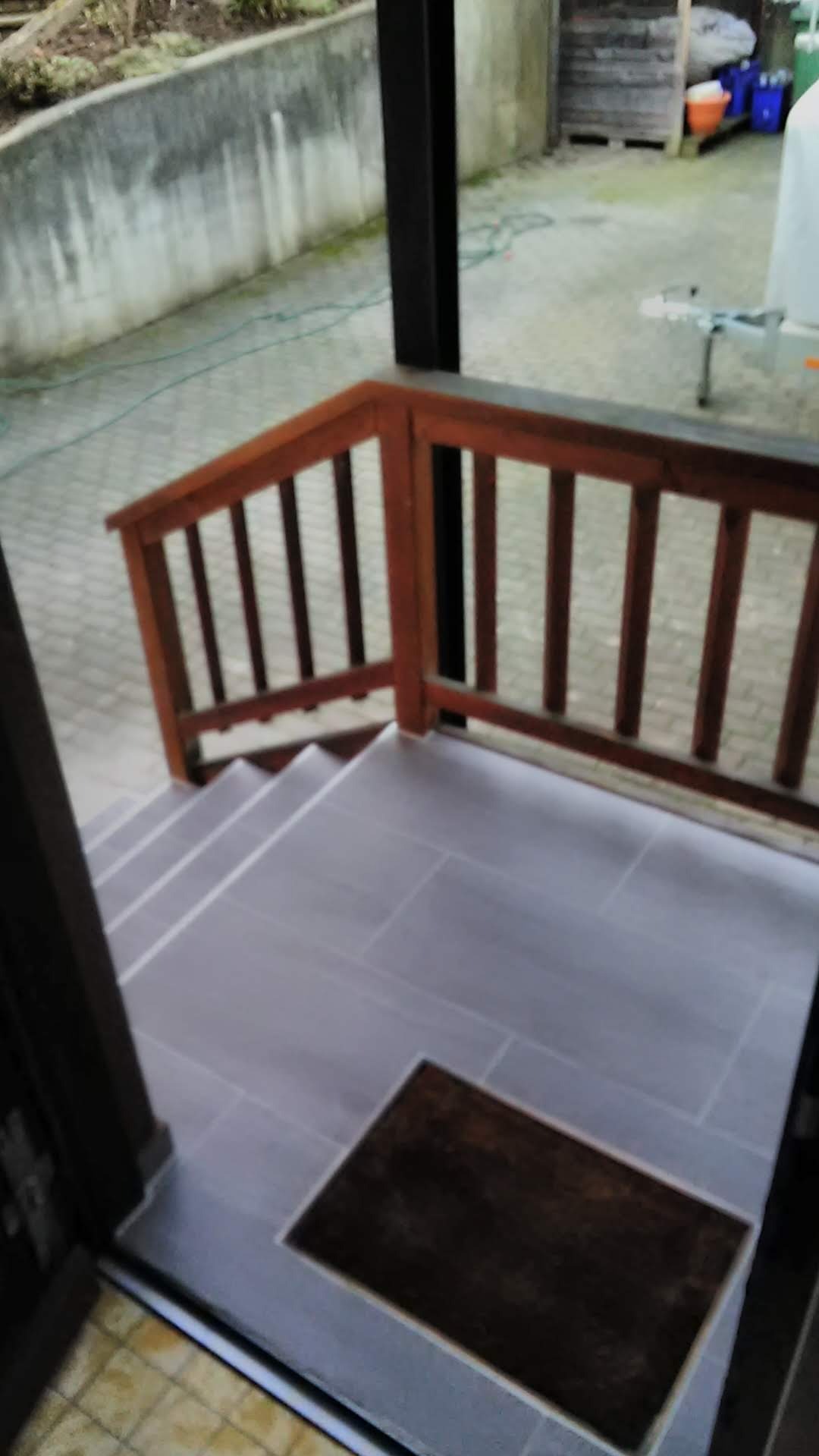 Wykończone płytkami schody wejściowe z drewnianą balustradą. Widok z góry, z brązową wycieraczką u dołu. Szare płytki, brązowe drewno, częściowo widoczne otoczenie.