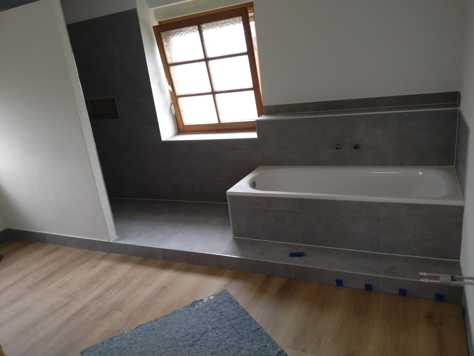 Wykończona łazienka z wanną w szarych płytkach, drewniane okno i podłoga. Widoczne detale wykończeniowe i fragment narzędzia.