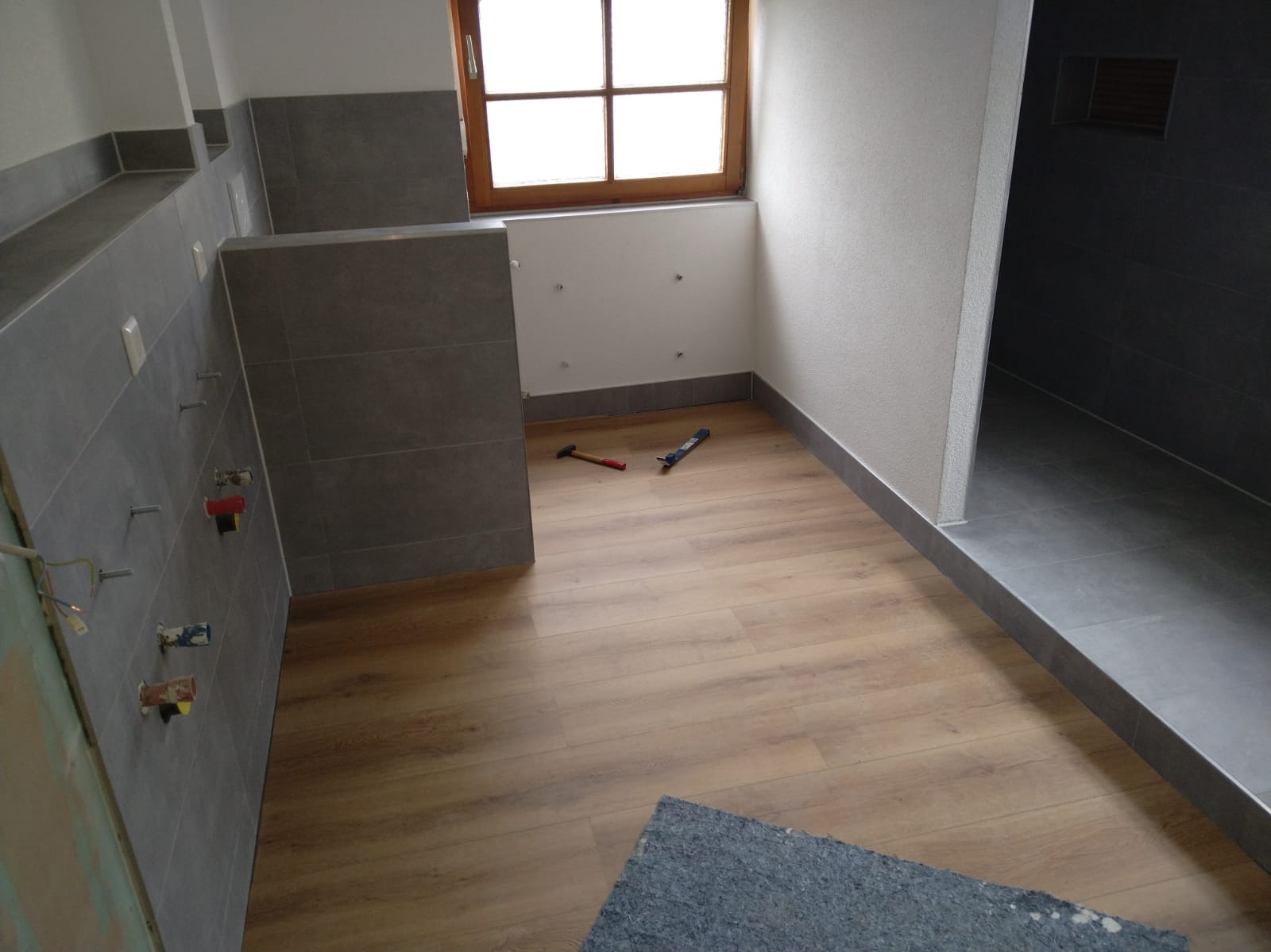 Wnętrze łazienki w trakcie prac wykończeniowych: położone płytki ścienne i podłogowe, widoczne narzędzia, okno z drewnianą ramą. Częściowo ułożone panele podłogowe.
