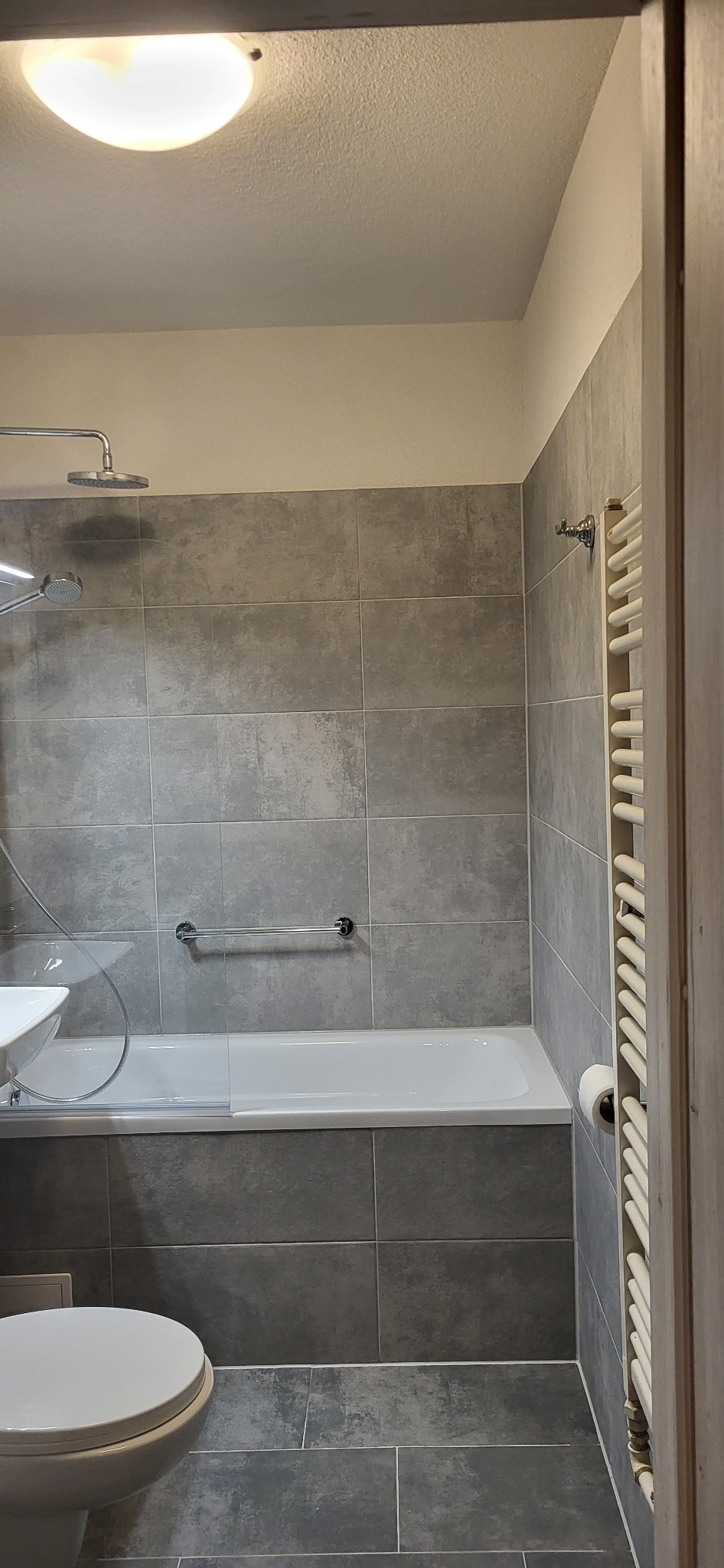 Wykończona łazienka w szarościach: płytki imitujące beton, biała wanna z parawanem, toaleta i grzejnik drabinkowy. Minimalistyczny design, oświetlenie sufitowe.