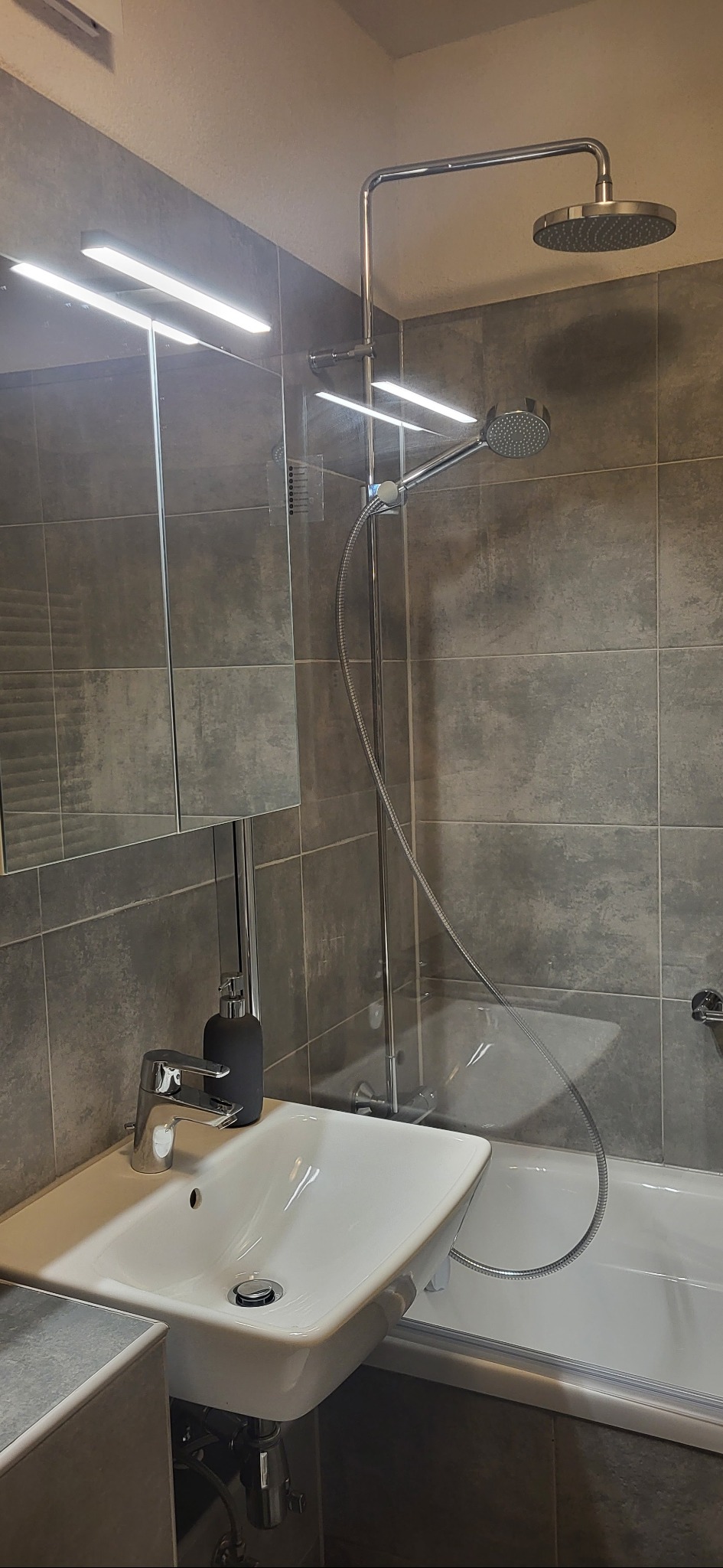 Nowoczesna łazienka z szarą glazurą, białą umywalką i wanną. Bateria umywalkowa chromowana, minimalistyczny design. Oświetlenie LED nad lustrem.